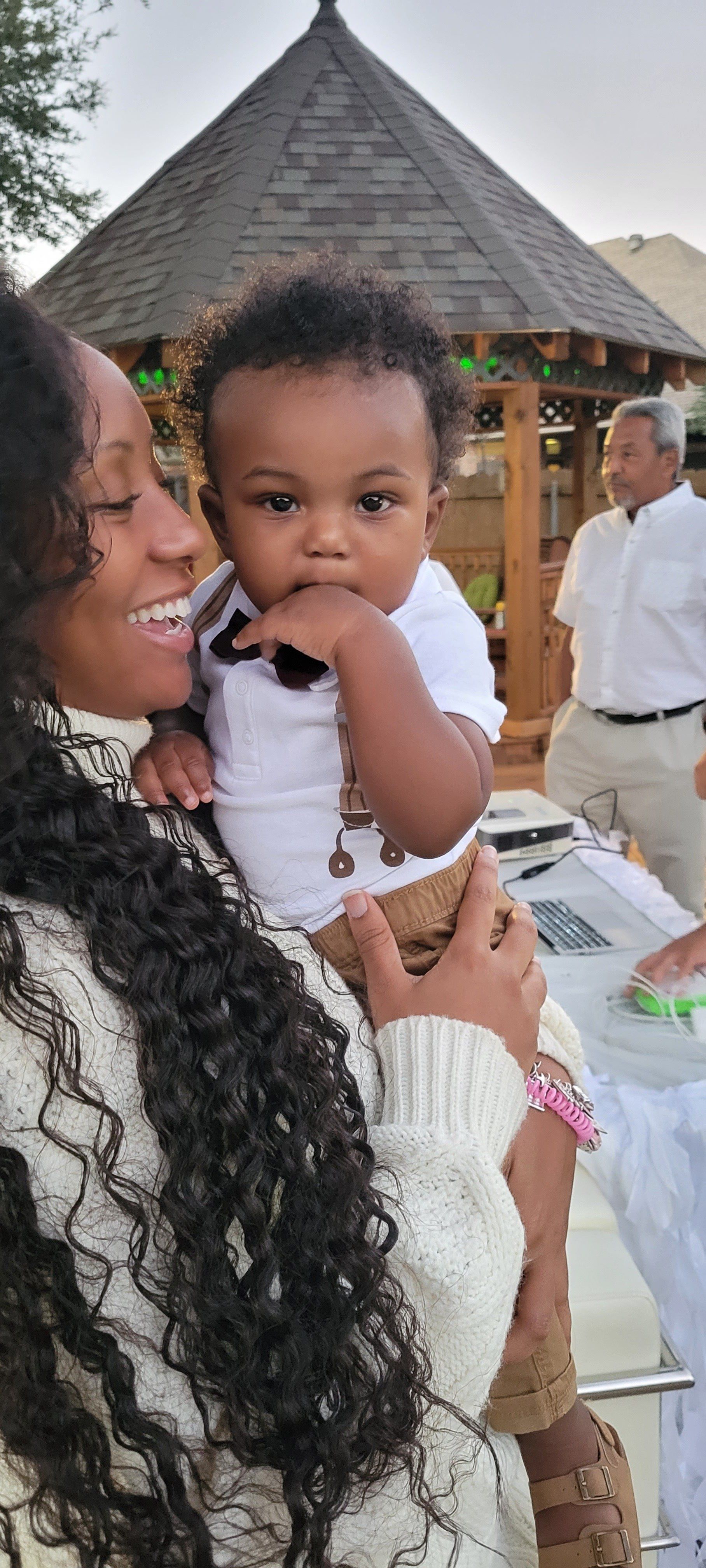 Woman holding a baby outside, smiling. Baby has hand in mouth. A man is in the background. Gazebo.