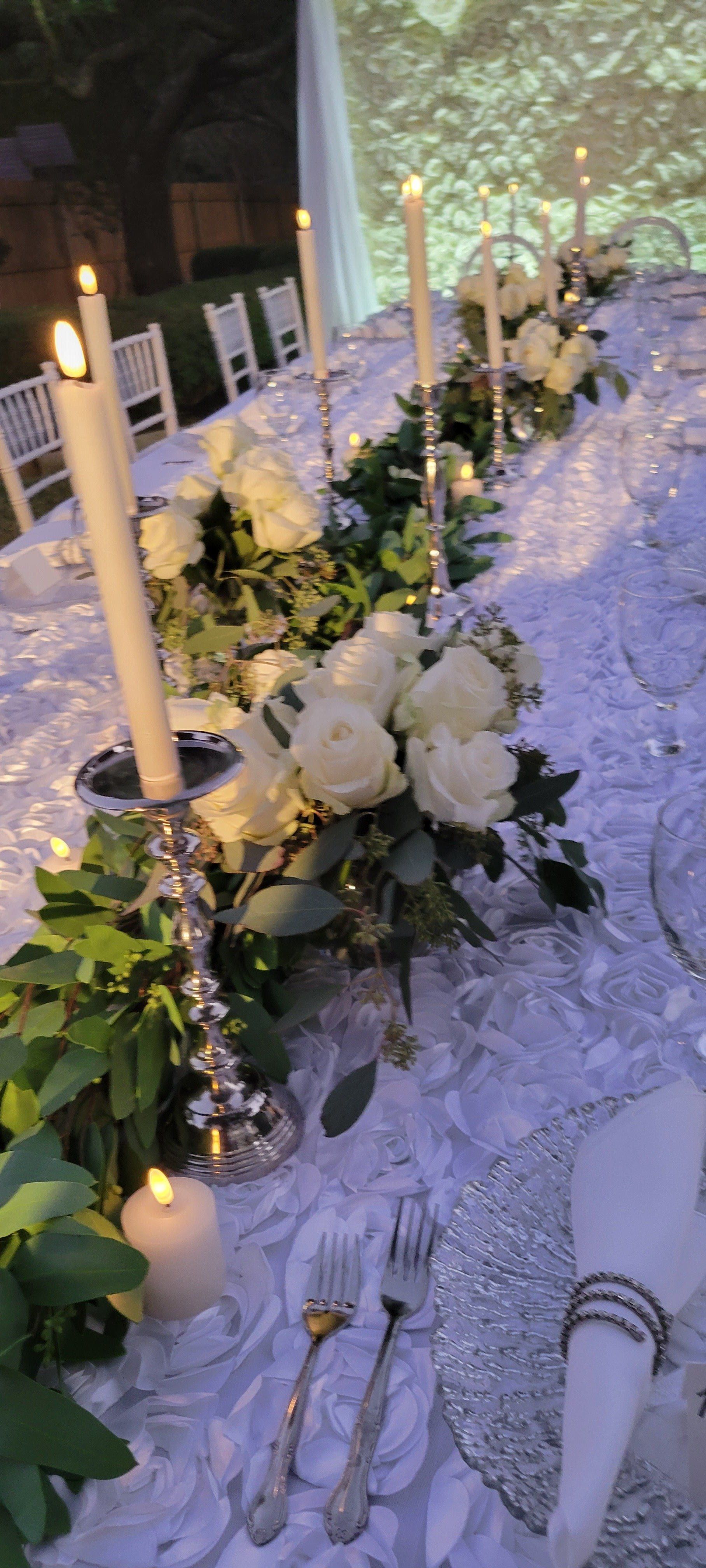 Table setting with white roses, candles, and greenery on a white tablecloth.