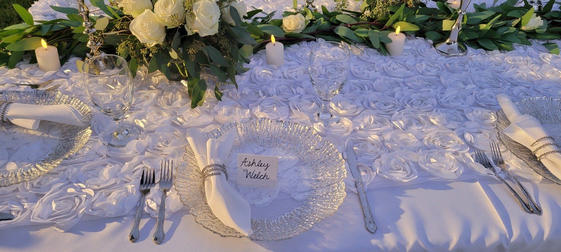 A white wedding table setting with flowers, candles, and a name card.