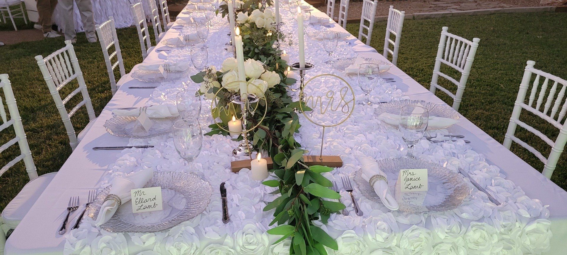 Long table set for an outdoor event, with white chairs and floral centerpieces.
