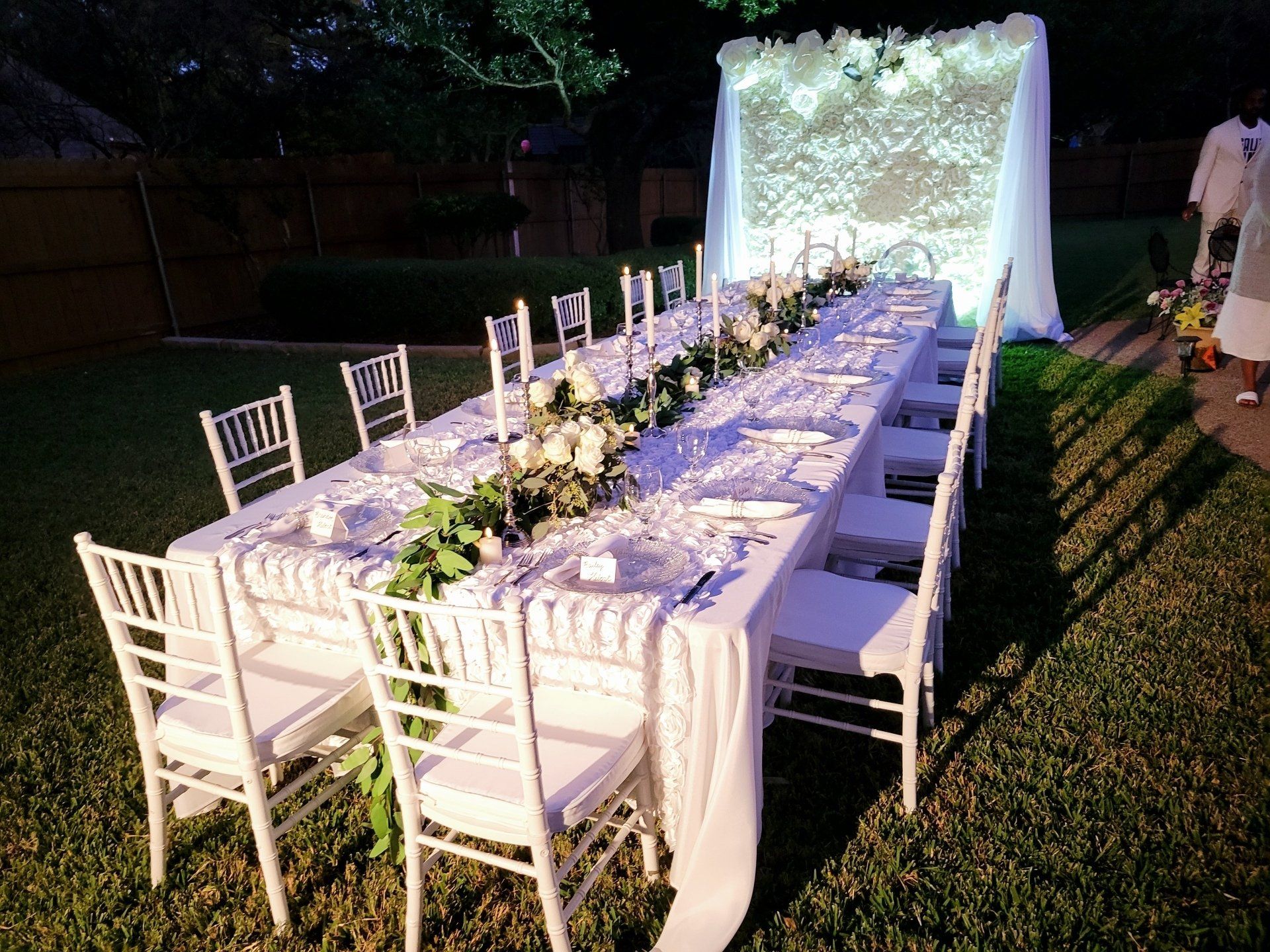 Long white table set for a formal outdoor event, with white chairs, flowers, and a floral backdrop.