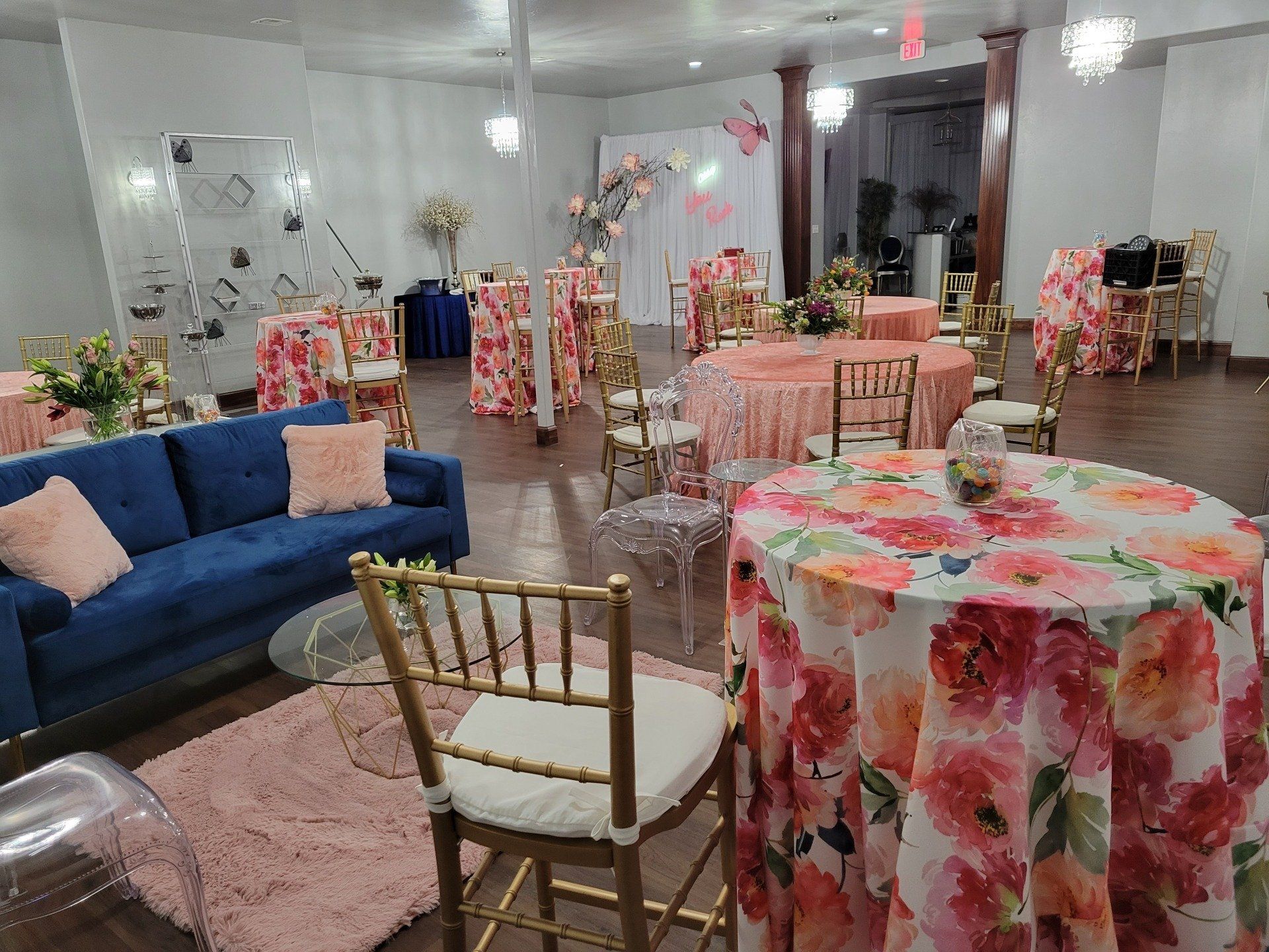 Event space with floral tablecloths, gold chairs, blue sofa, and decorations.
