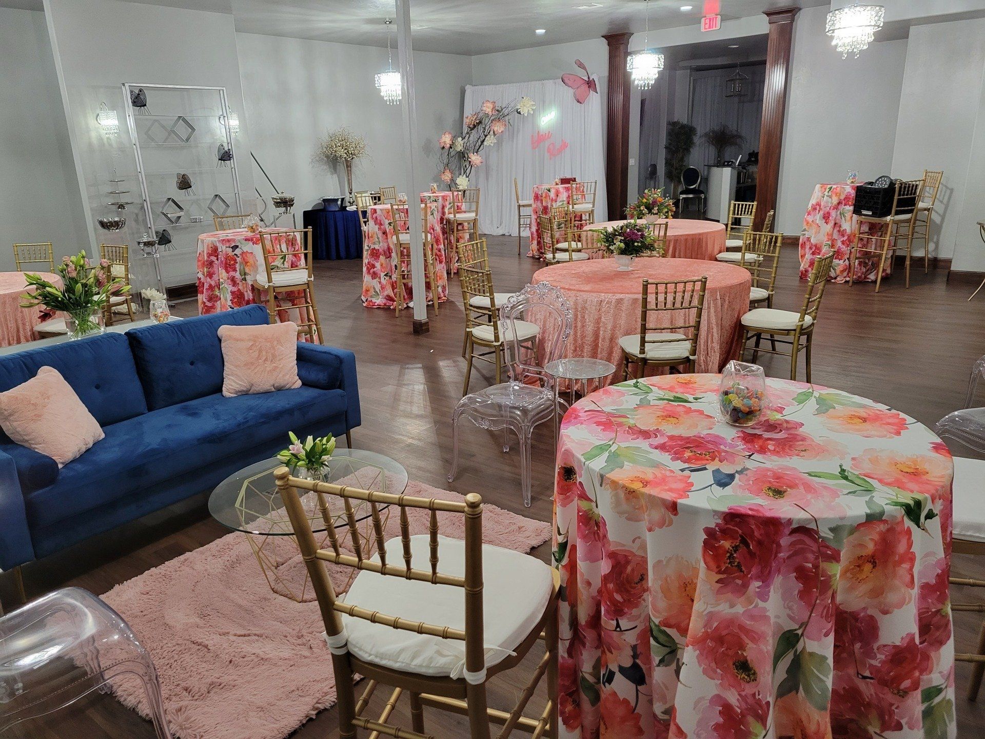 Party venue with floral tablecloths, gold chairs, and a blue sofa.