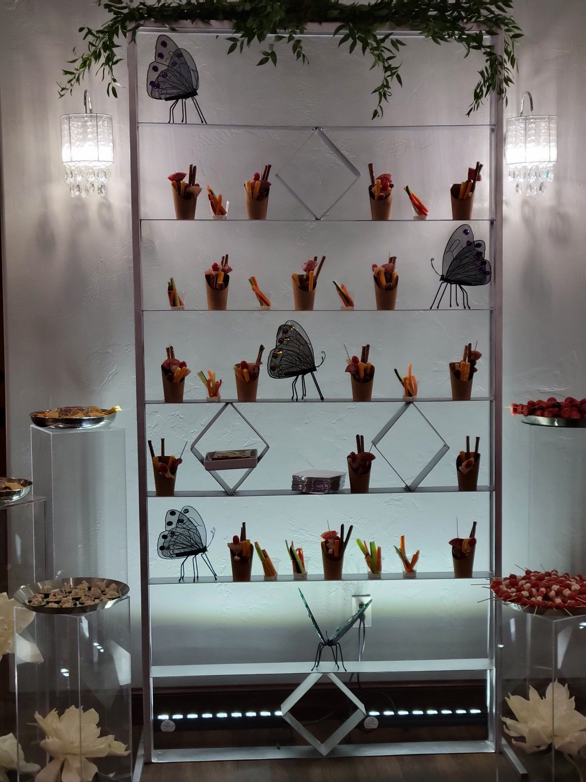 Dessert display with tiered shelves, treats in cups, illuminated. Butterflies and greenery decorate.