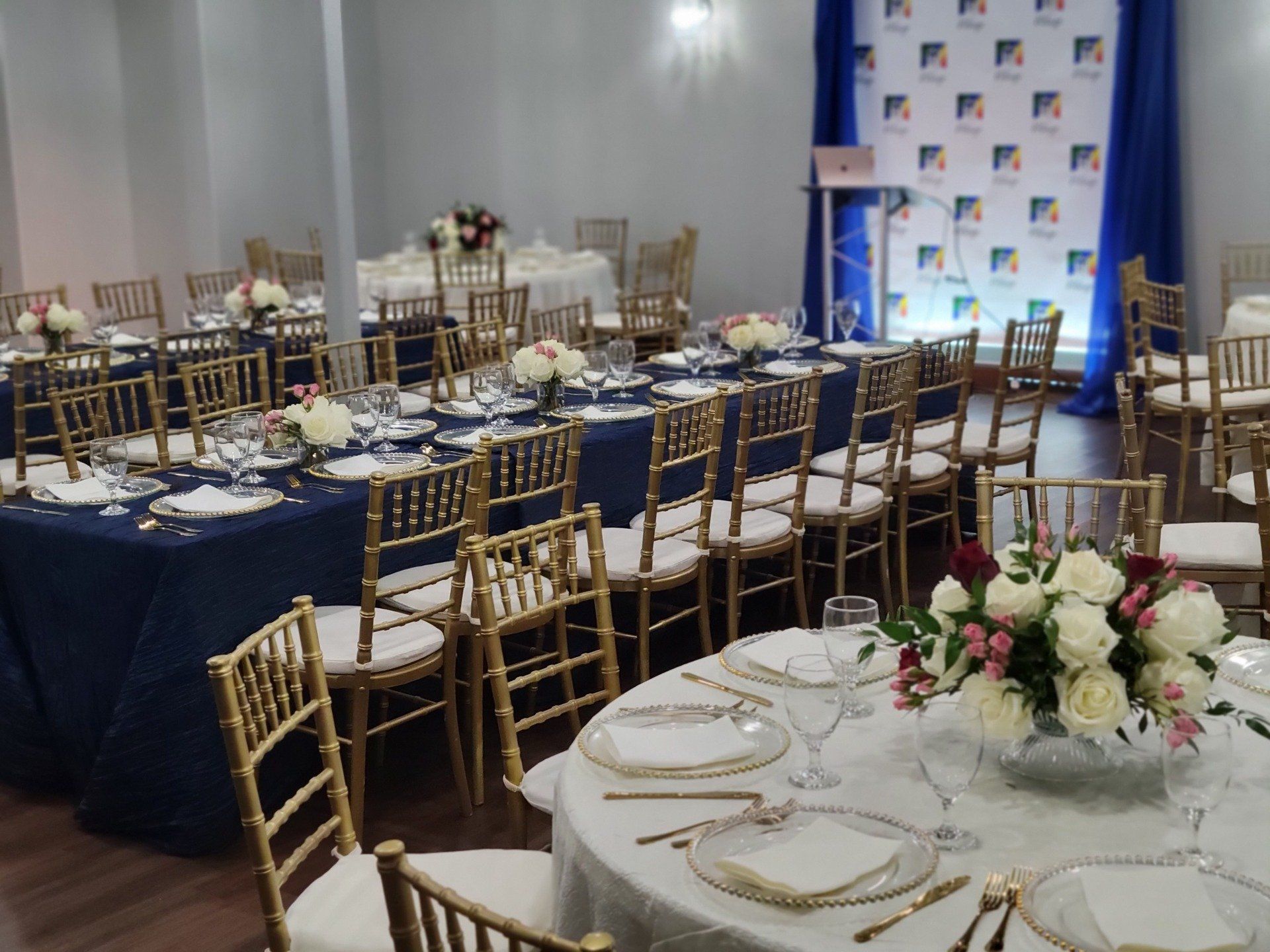 Event hall with tables set for a meal. Gold chairs, blue and white linens, floral centerpieces.