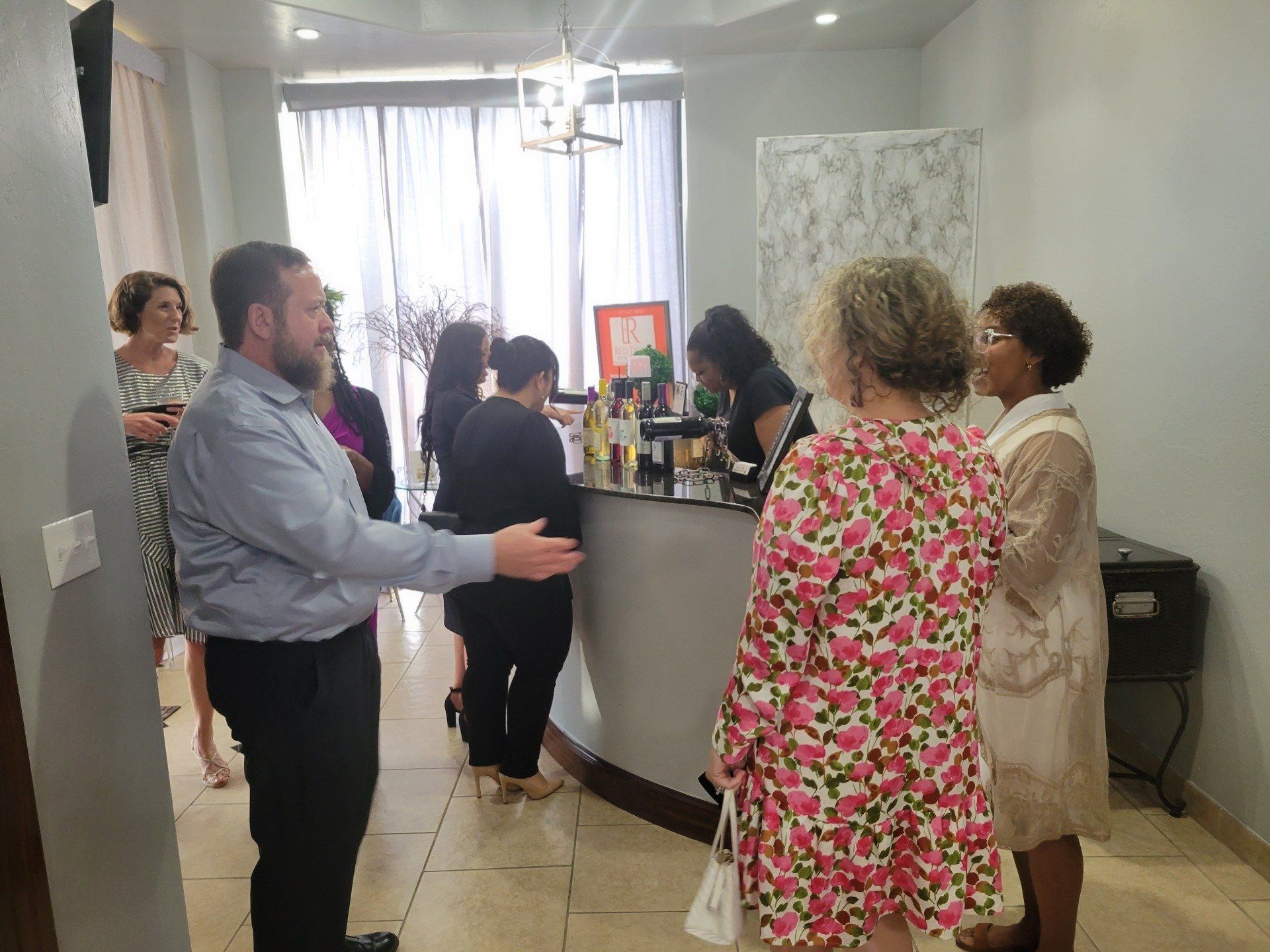 People at an event, man gesturing, standing near a counter with others, in a room with a window.
