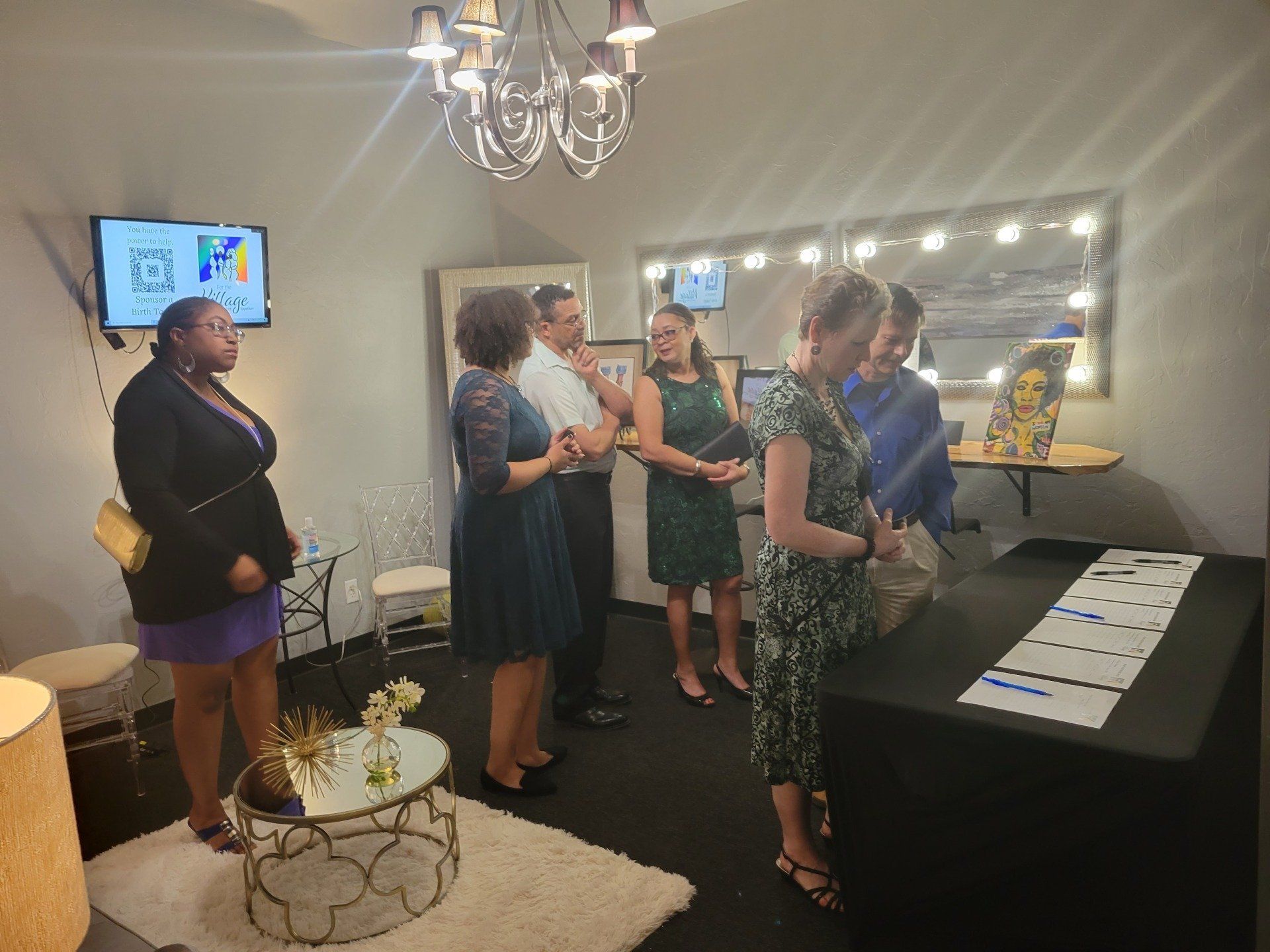 People in a room looking at artwork and a table, some are talking. There's a lighted mirror and a small round table.