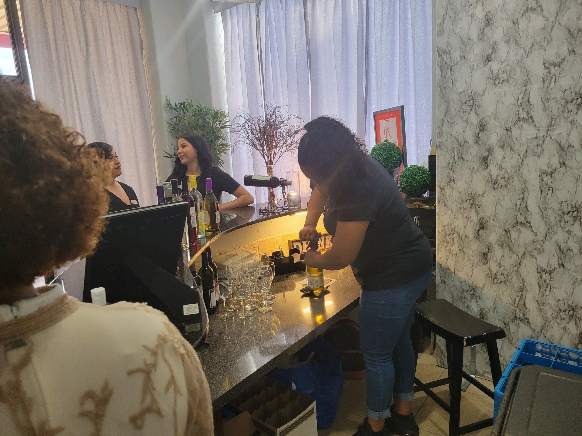 Woman behind bar opening a bottle, others nearby. Interior with light and decorative elements.