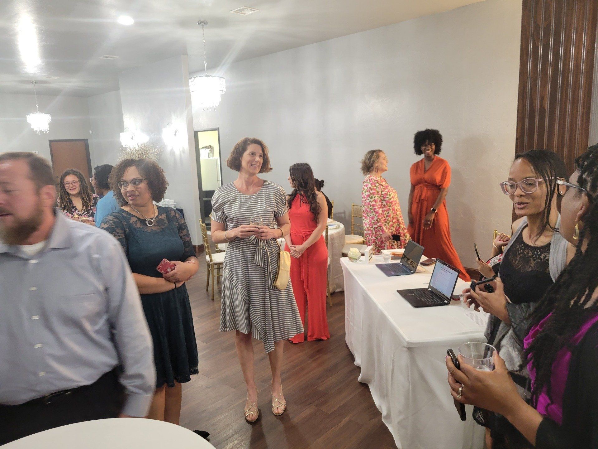 People at an event in a well-lit room. Some are standing near a table with laptops, others are conversing.
