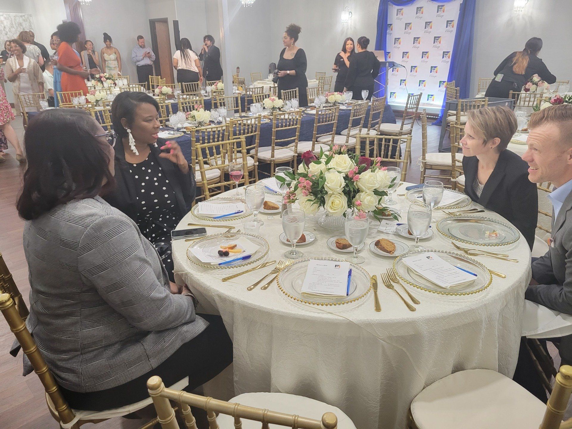 People seated around a decorated table at an event. Others socialize in the background.