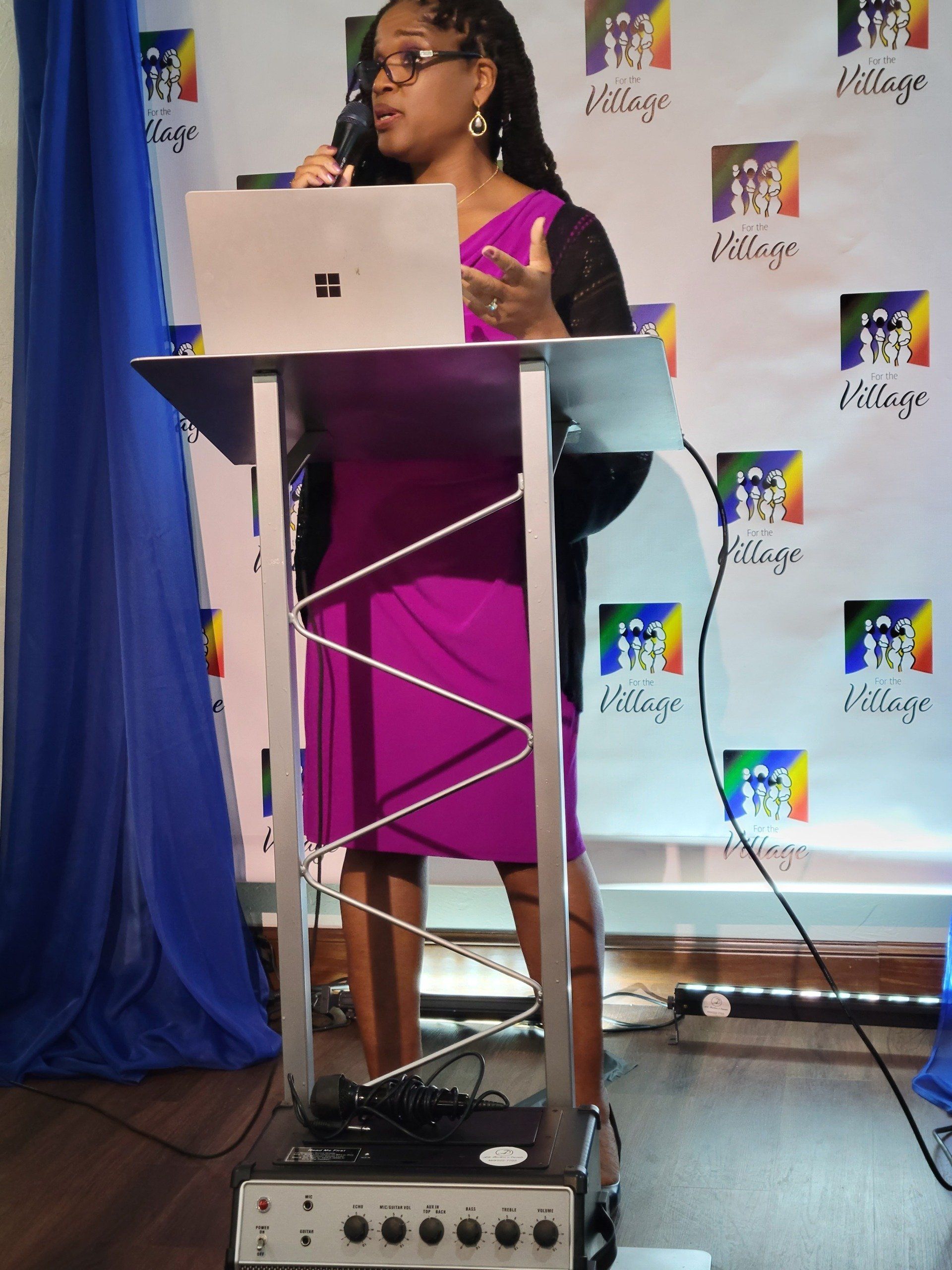 Woman speaking at a lectern, presenting with a laptop. Backdrop with logos.
