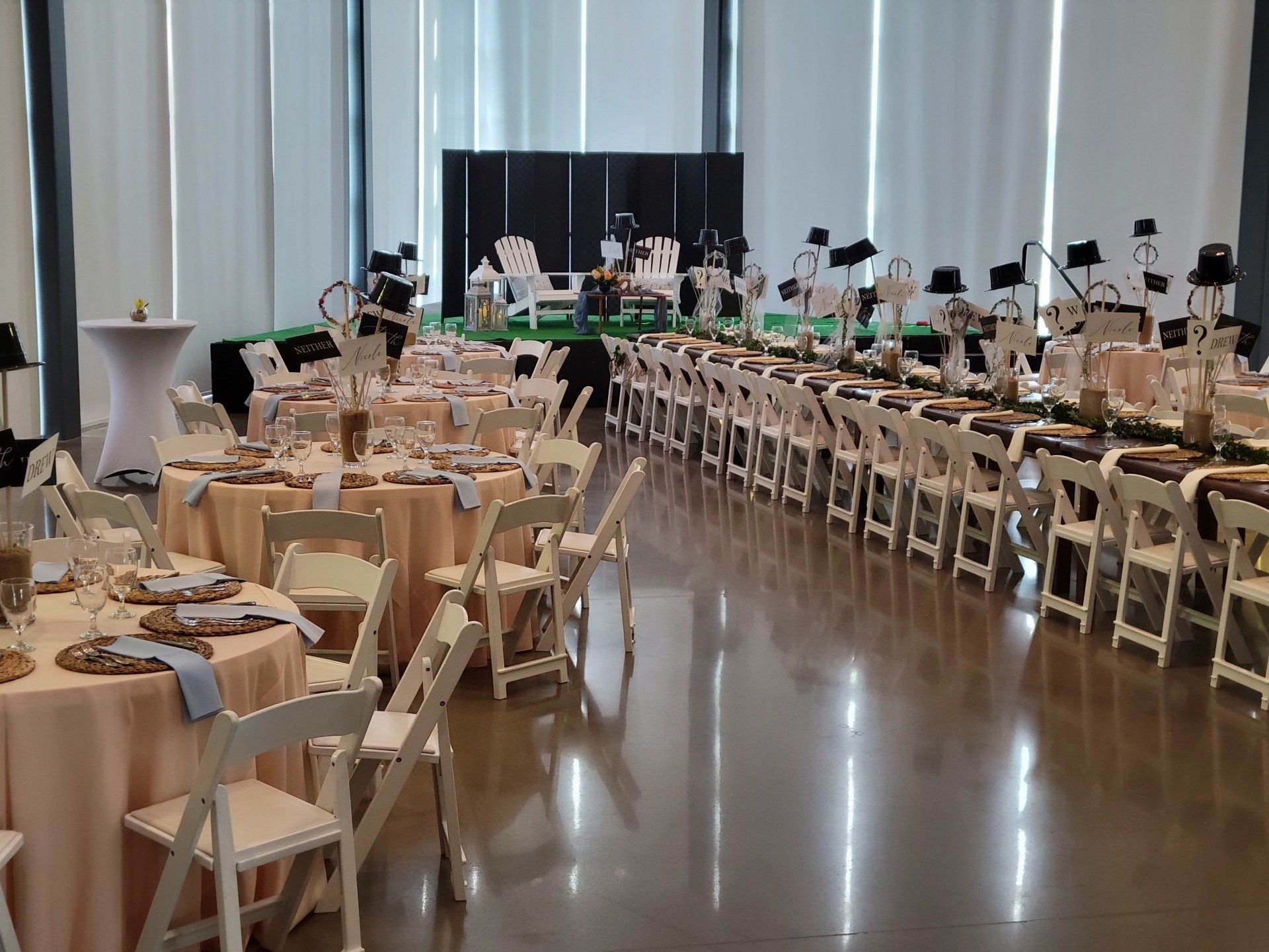 Wedding reception setup: tables with blush linens, white chairs, stage with instruments, green runner.