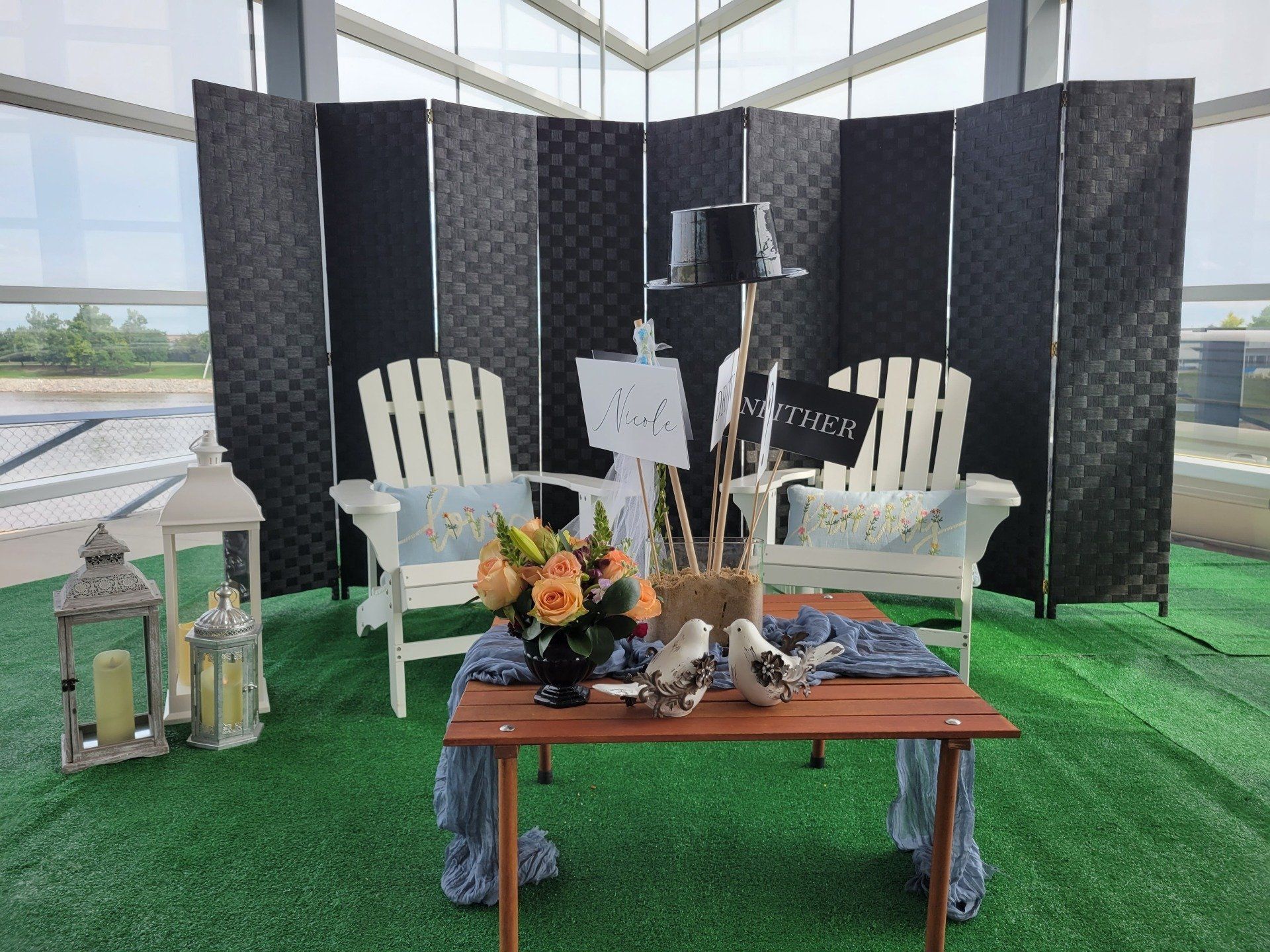 An outdoor wedding setup: two white chairs, wooden table with floral arrangement and black screen backdrop.
