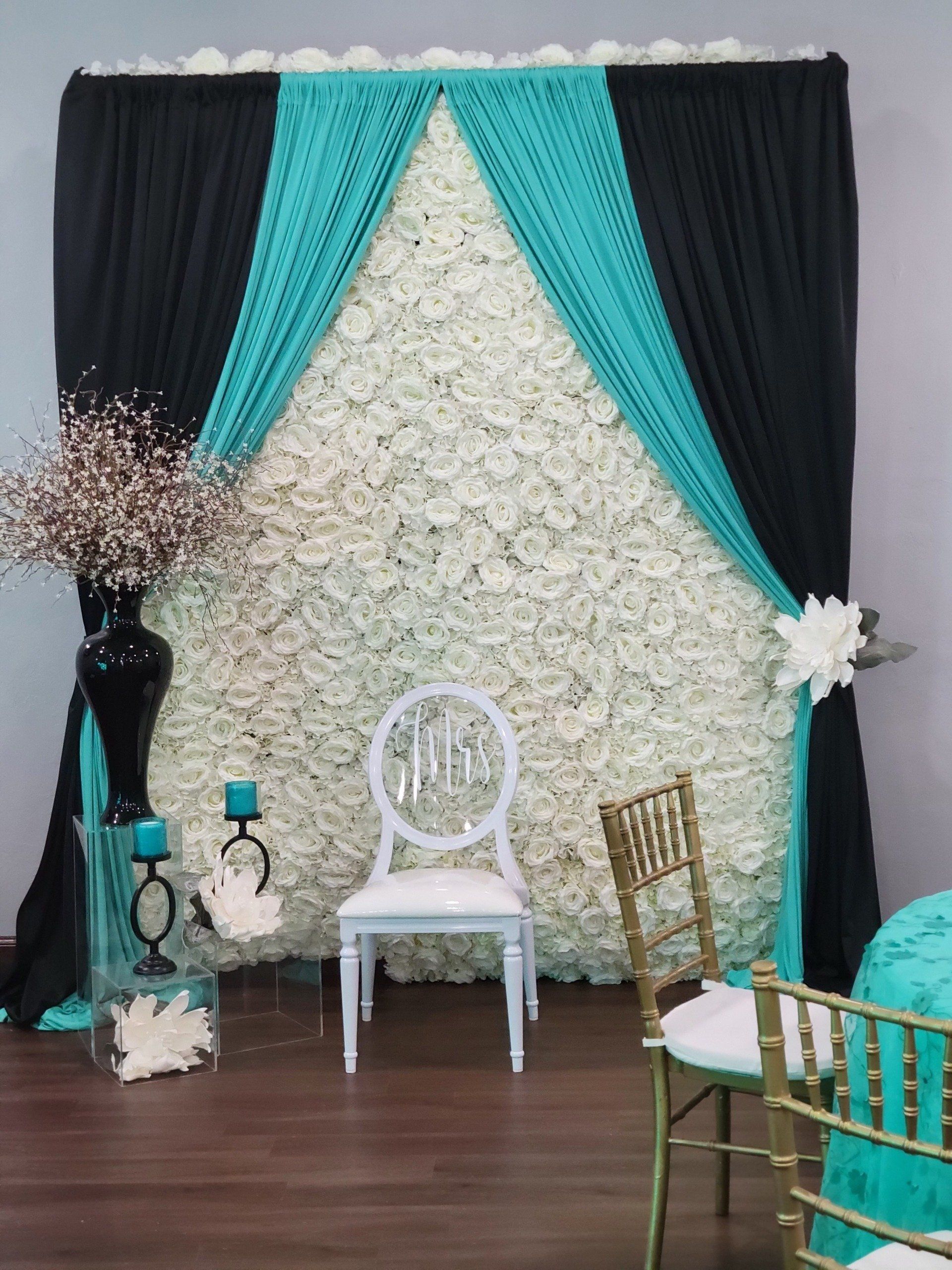 Wedding backdrop with white rose wall, teal and black drapes, white and gold chairs, decorative elements.