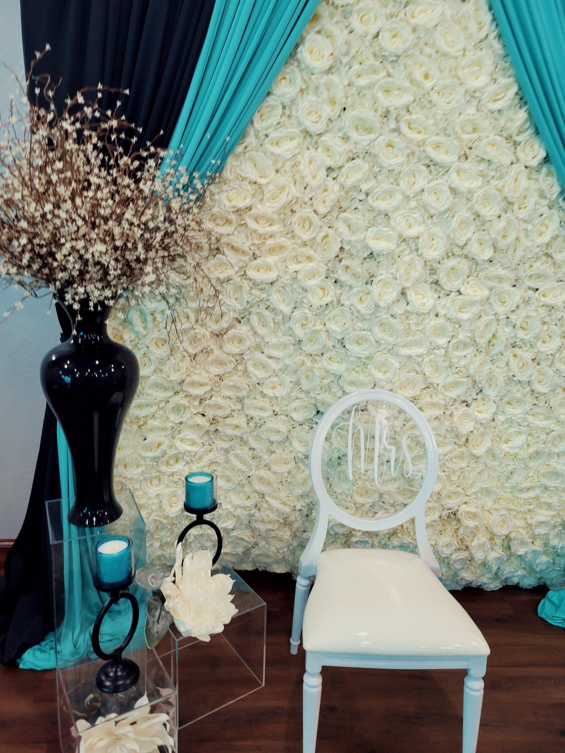 Wedding backdrop with white flower wall, teal accents, and a white chair.