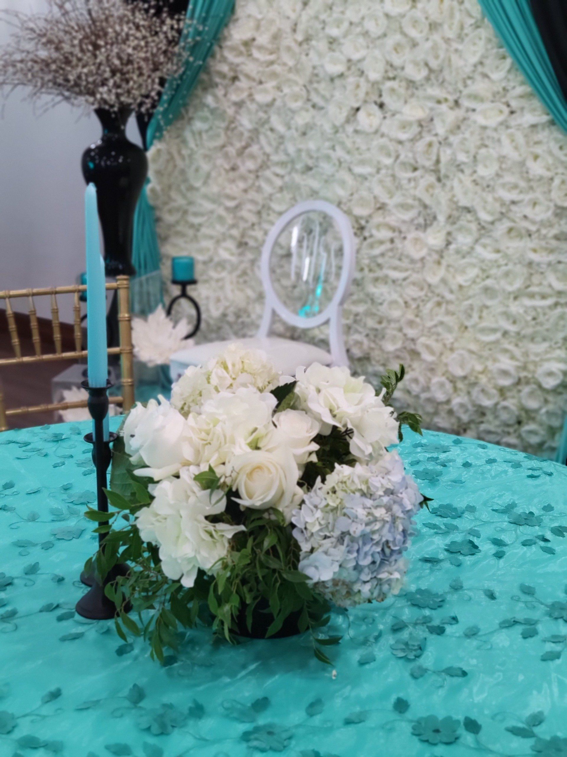 Table setting with white flowers, blue candles, and white chair against flower wall.