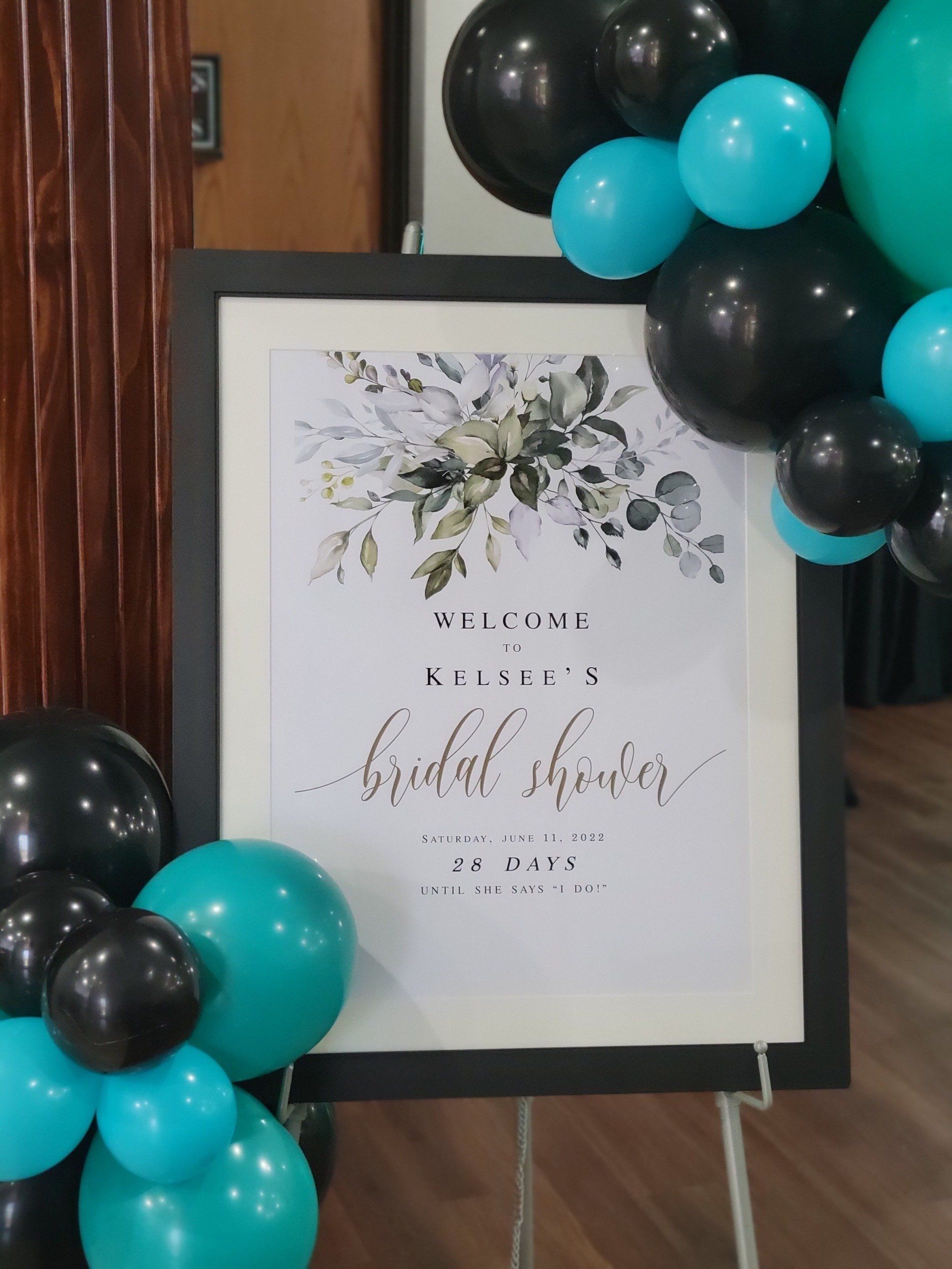 Bridal shower sign with floral design, black frame, turquoise and black balloons.