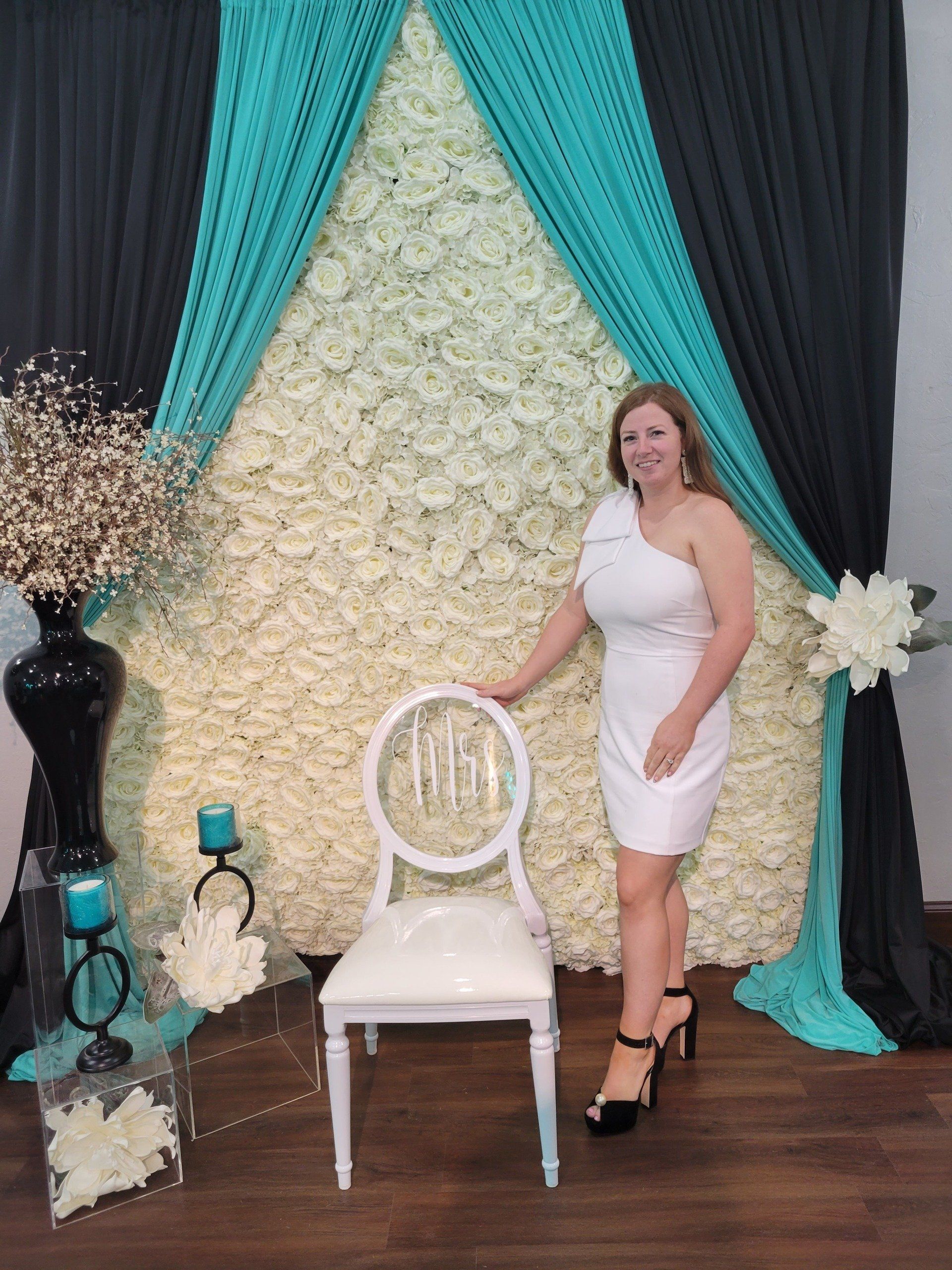 Woman in white dress posing by a floral backdrop, with turquoise and black drapes, chair, and decorative accents.