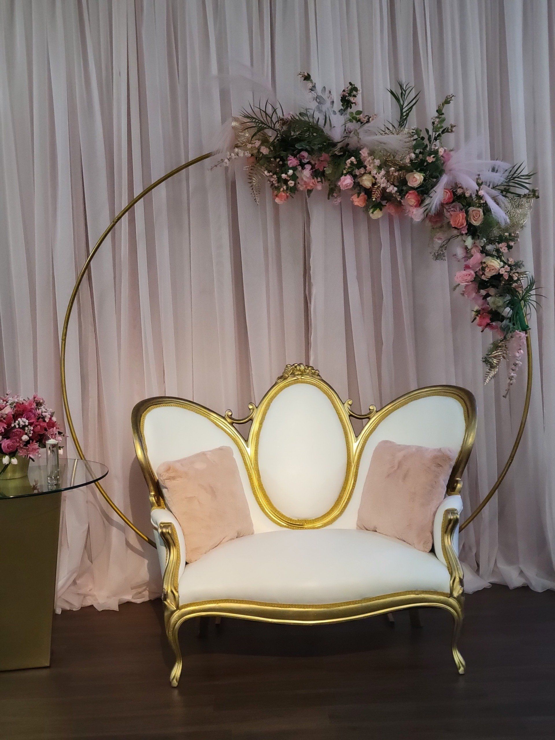 White and gold ornate loveseat with pink pillows set before a floral-adorned gold ring and draped pink fabric.