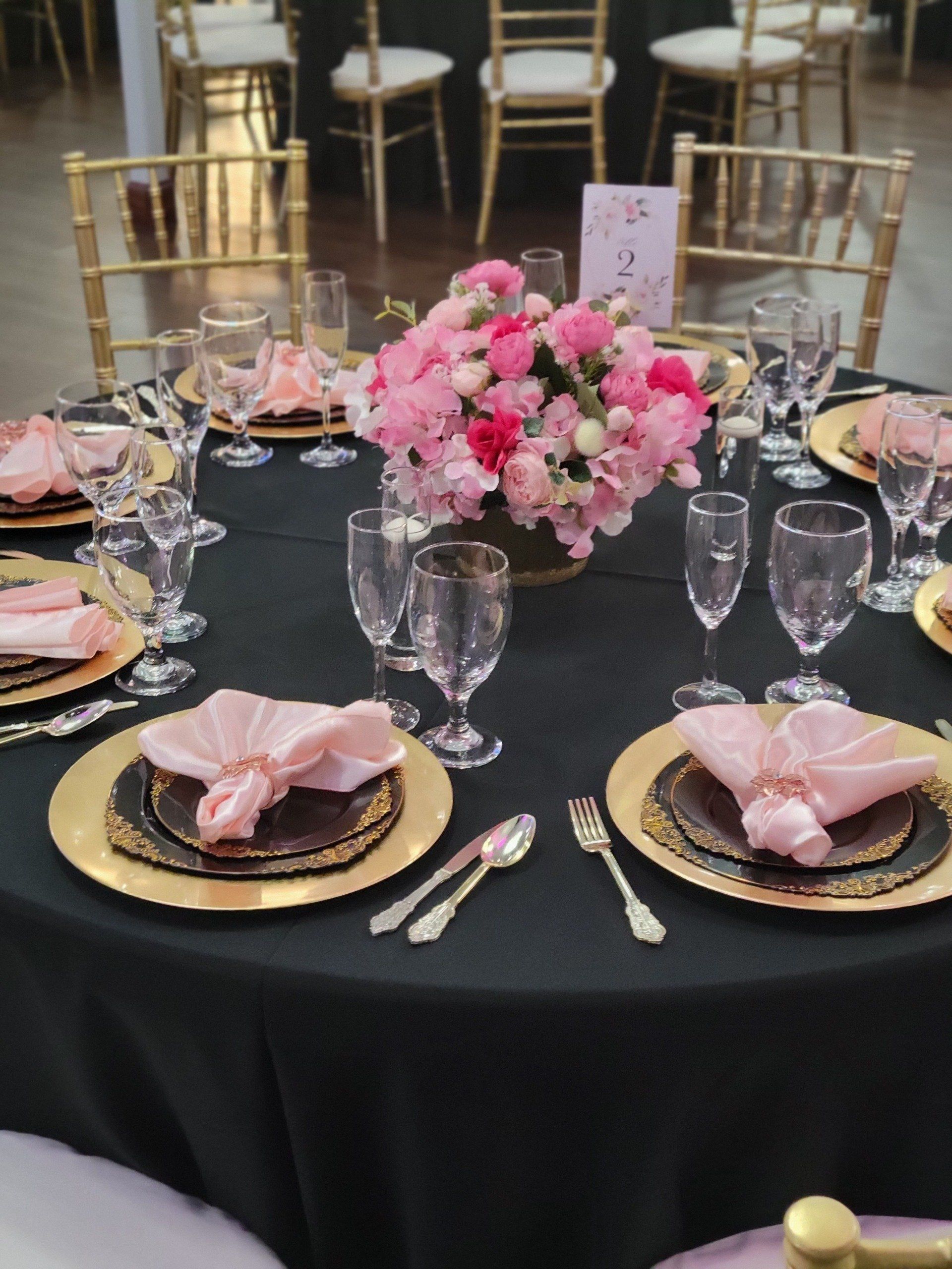 Elegant table setting with black tablecloth, gold chargers, pink napkins, and floral centerpiece.