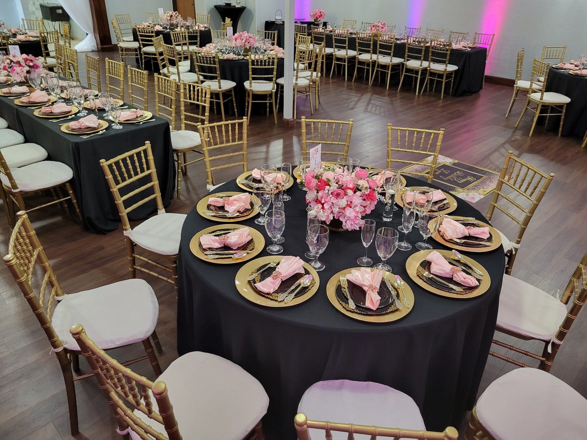 Wedding reception: Tables with gold chairs, black tablecloths, pink decor. Wooden floor, overhead lighting.