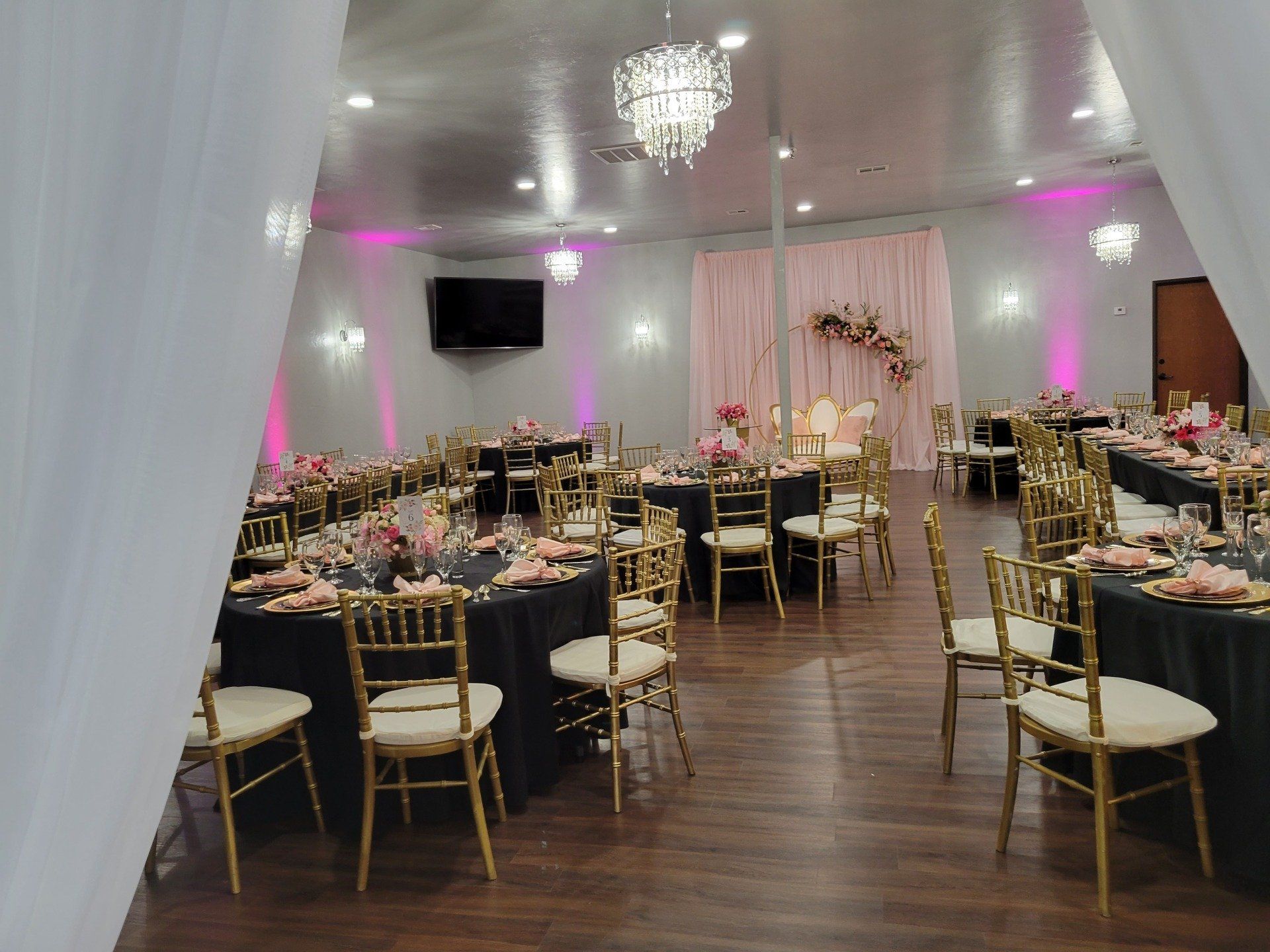 Banquet hall with black tablecloths, gold chairs, pink uplighting, and a floral backdrop.