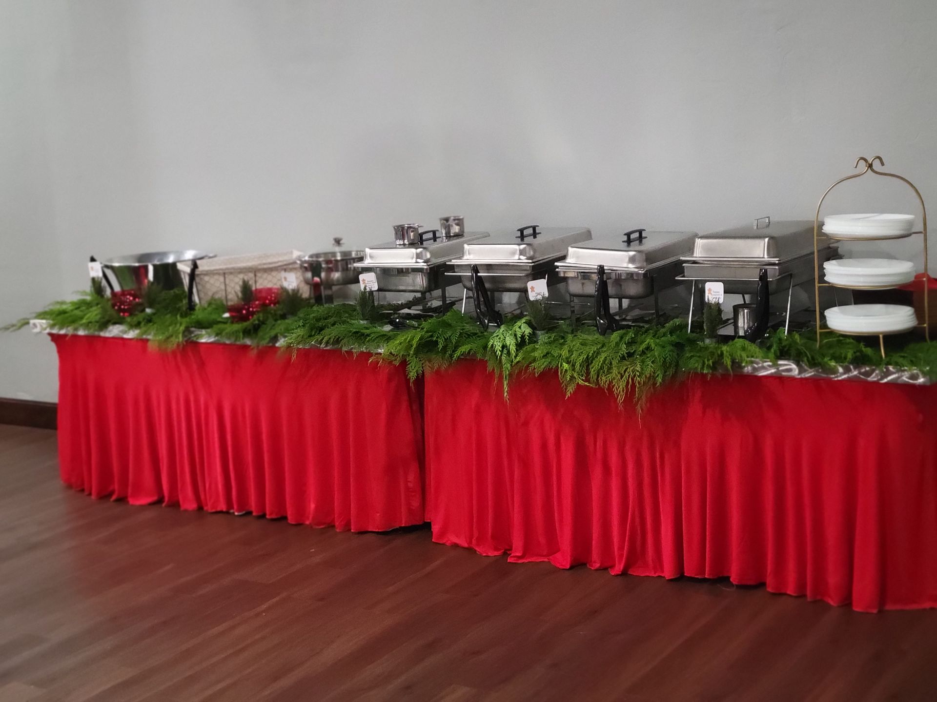 Red buffet tables with chafing dishes, greenery, and serving platters set against a white wall and wood floor.