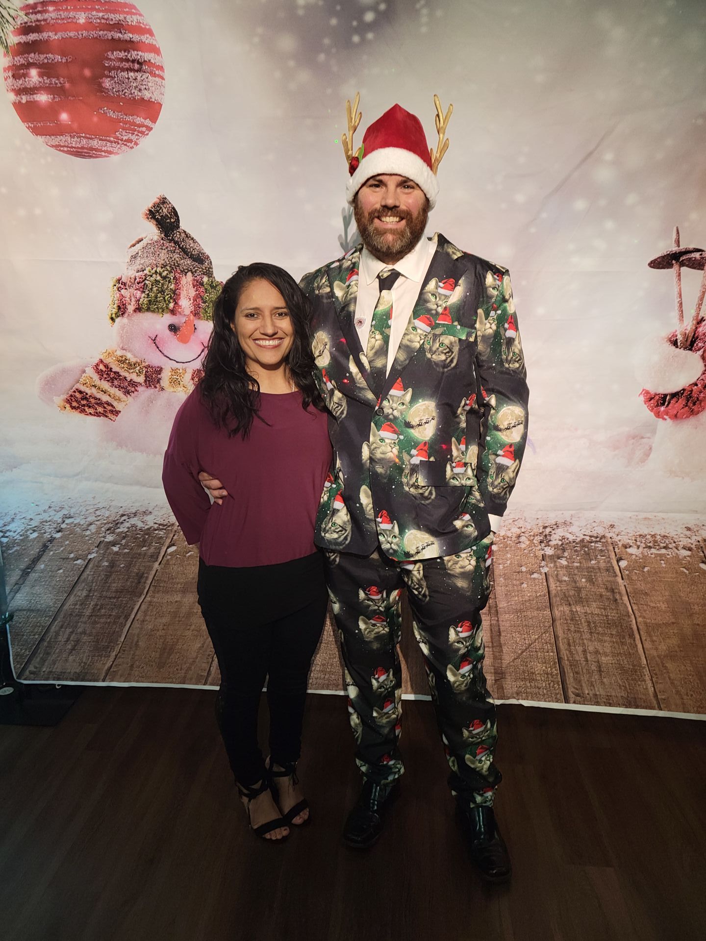 Woman and man posing in front of Christmas backdrop. Man wears a festive suit and antlers. Woman has her arm around the man.