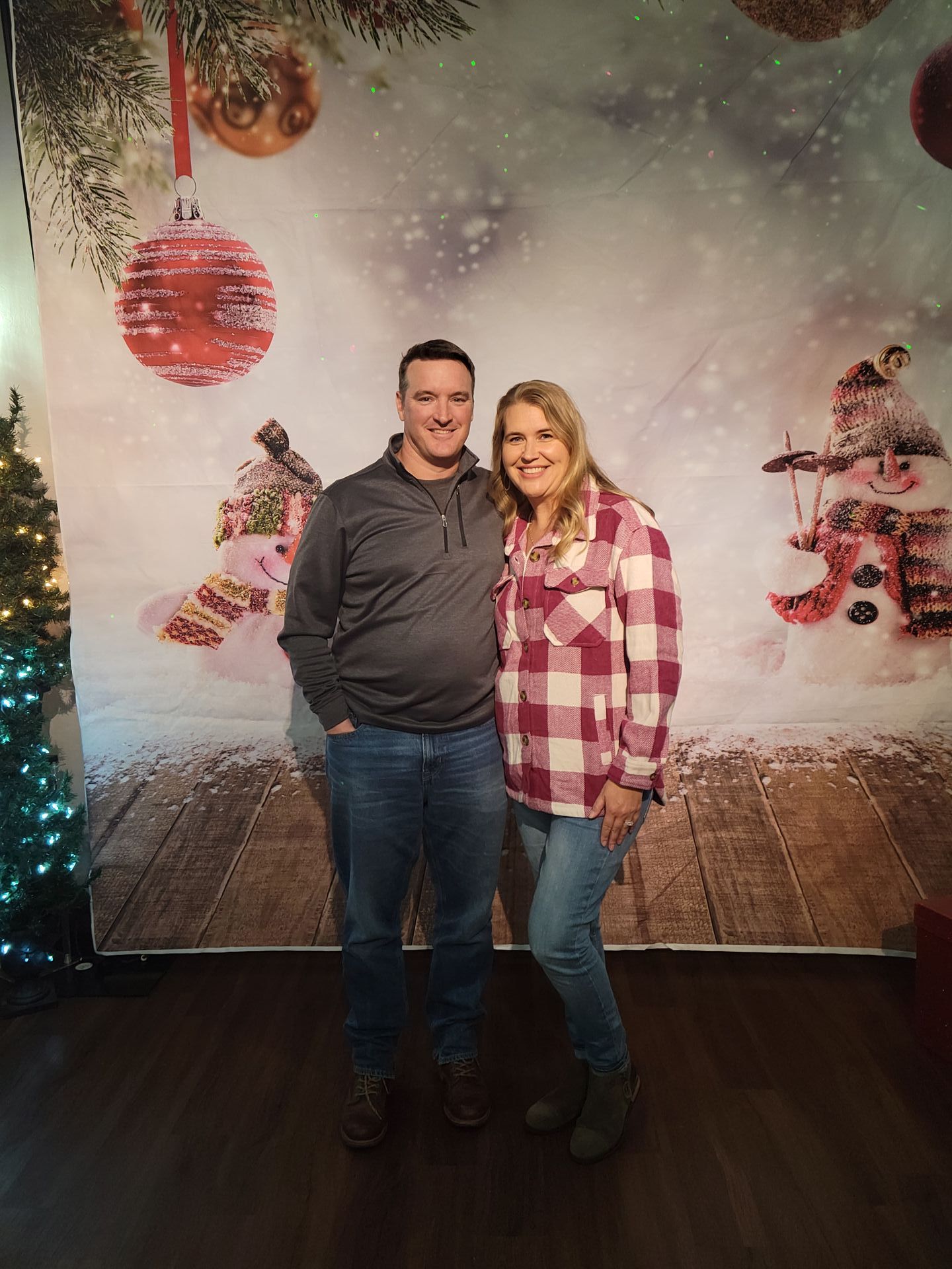 Couple poses in front of a Christmas backdrop with ornaments and snowmen. Woman wears a red plaid shirt.