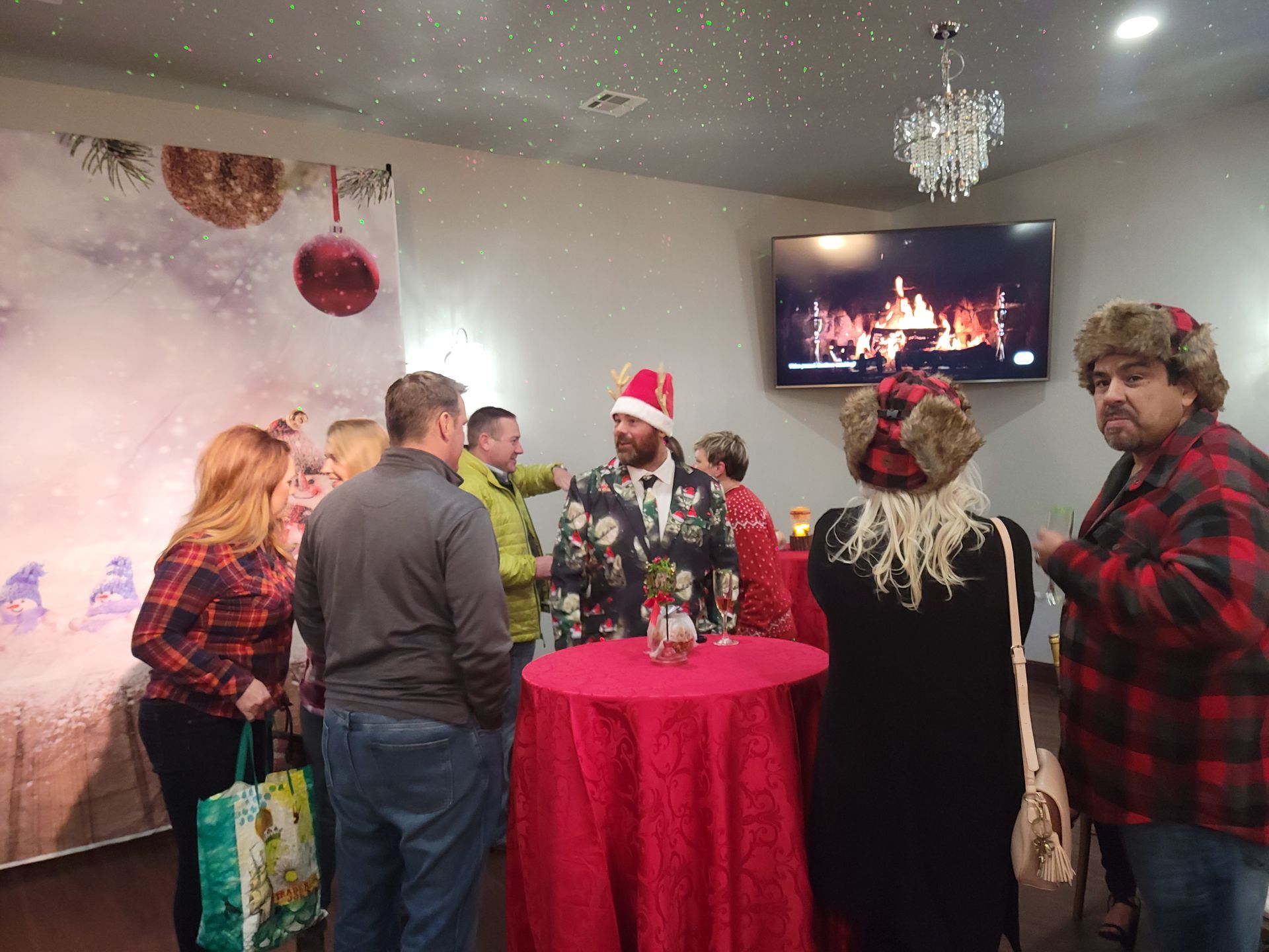 People gathered at a Christmas party. A man in a festive suit and hat stands by a red table.