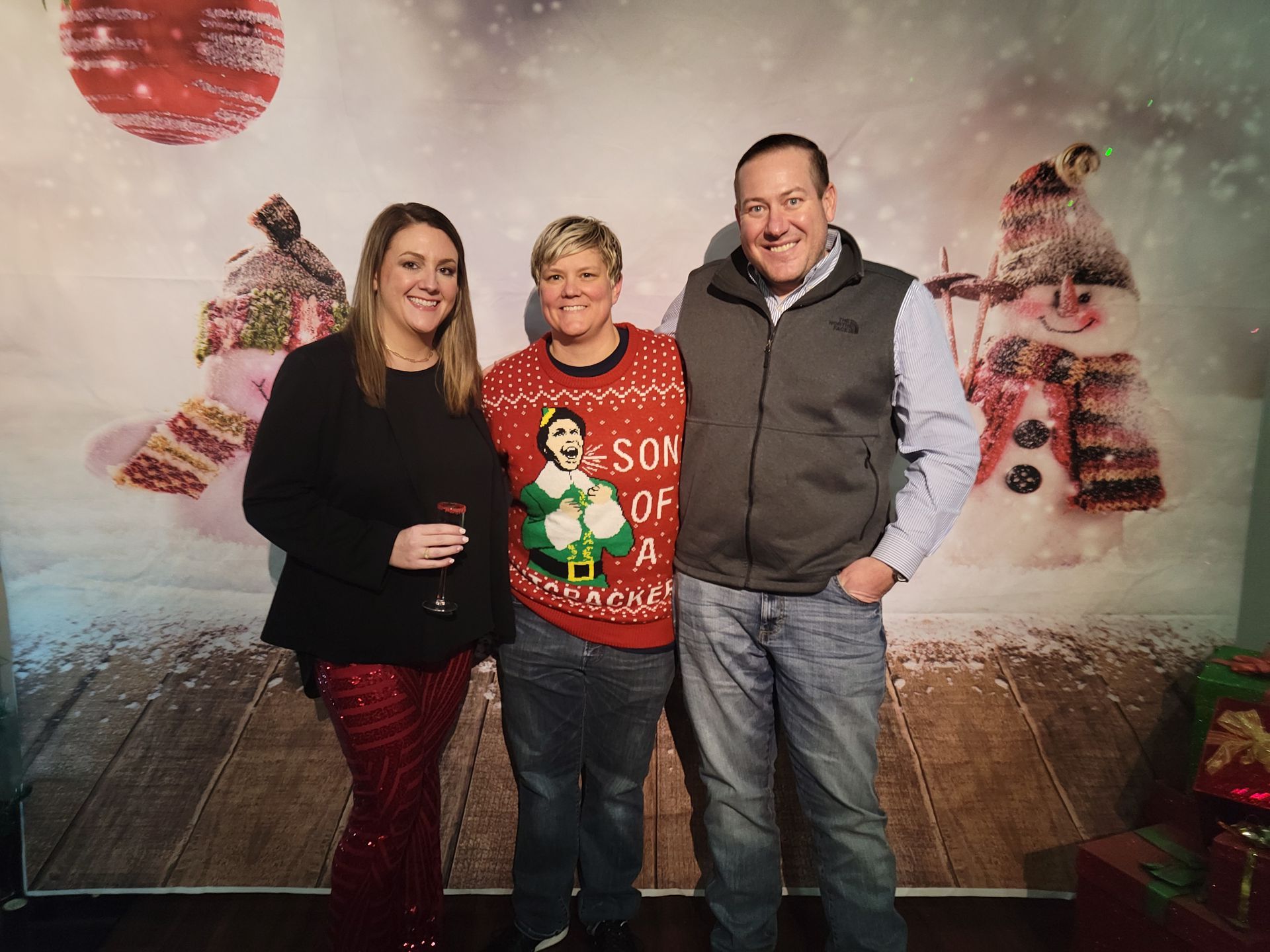 Three people posing in front of a Christmas backdrop. Woman in red sequined pants and person in a festive sweater.