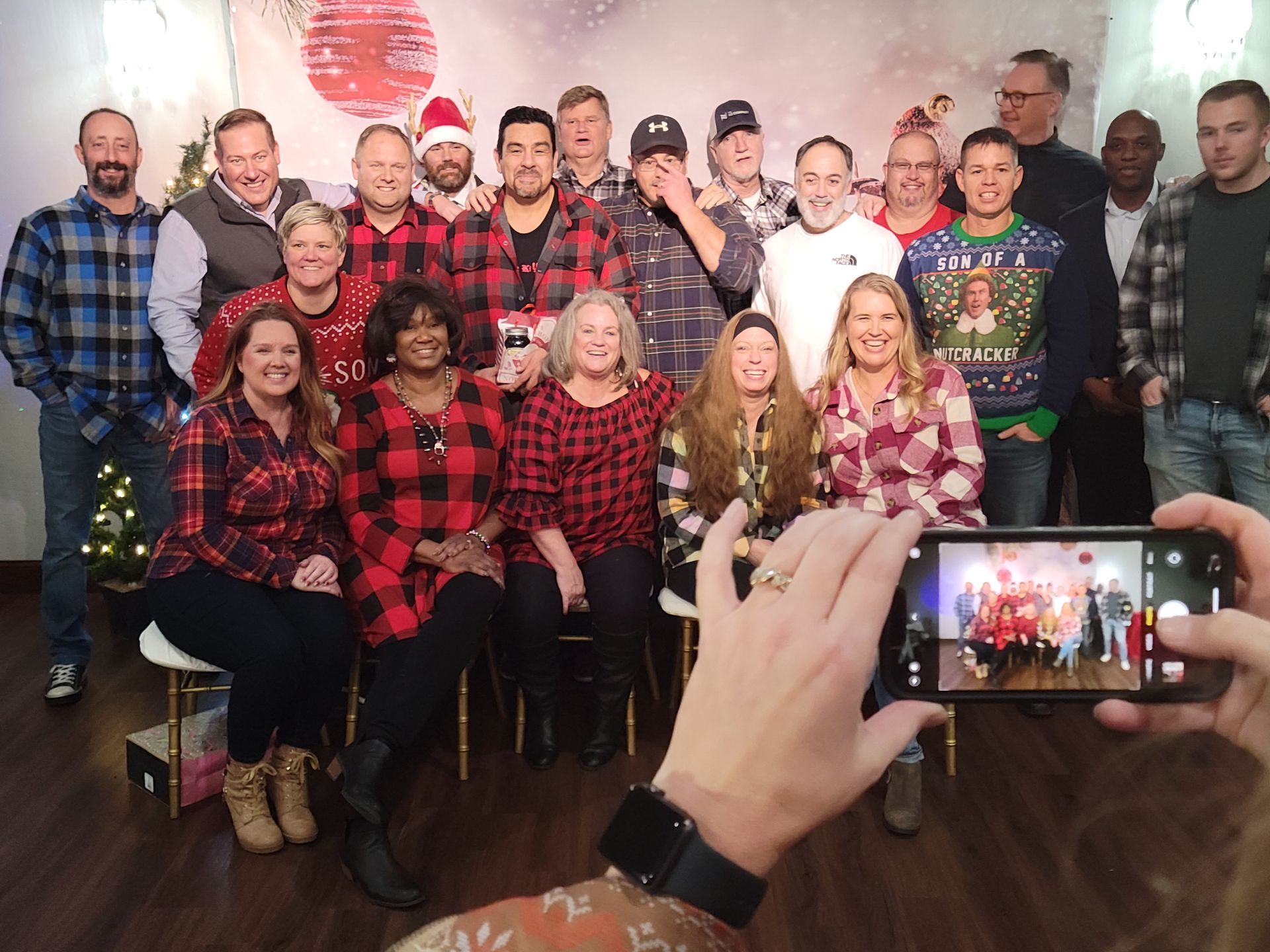 Group photo in front of a holiday backdrop, people wearing red plaid, some with sweaters.