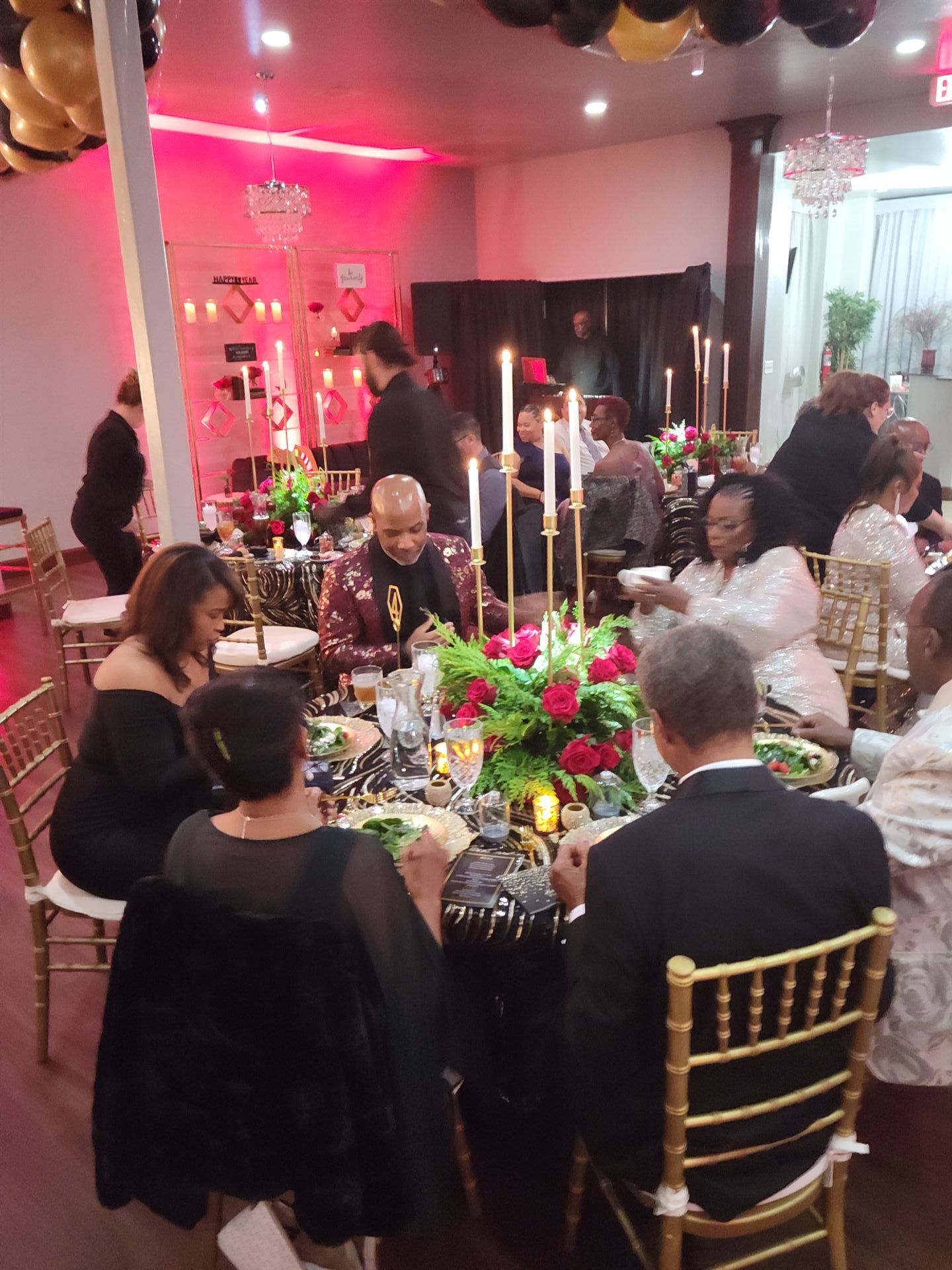 Guests seated at a formal dinner, tables set with centerpieces, lit candles. Black and gold decor, red accent.