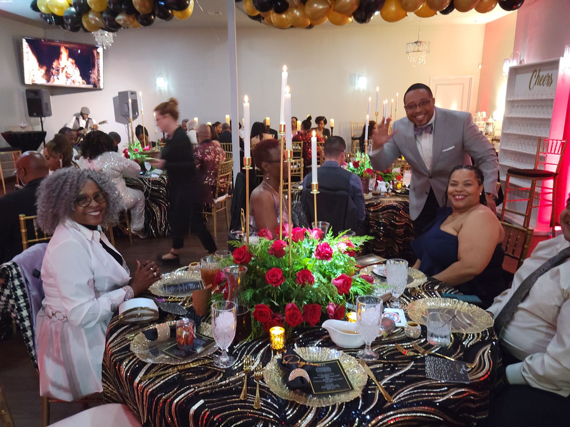 People at a formal event, seated at tables with centerpieces, candles, and gold accents. One man waves.