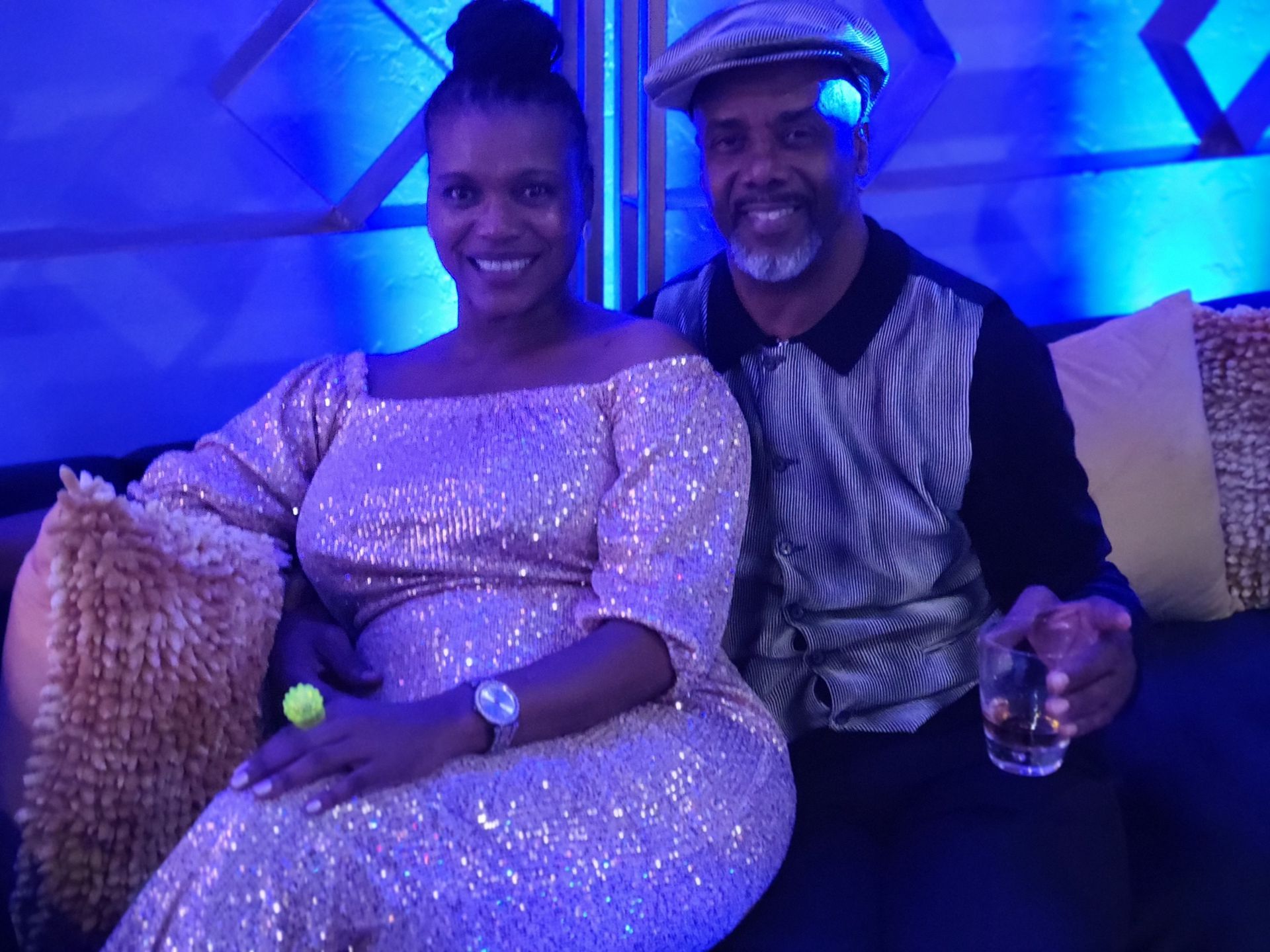 Woman in sequined dress and man with cap sitting, smiling. They are in a blue-lit room.