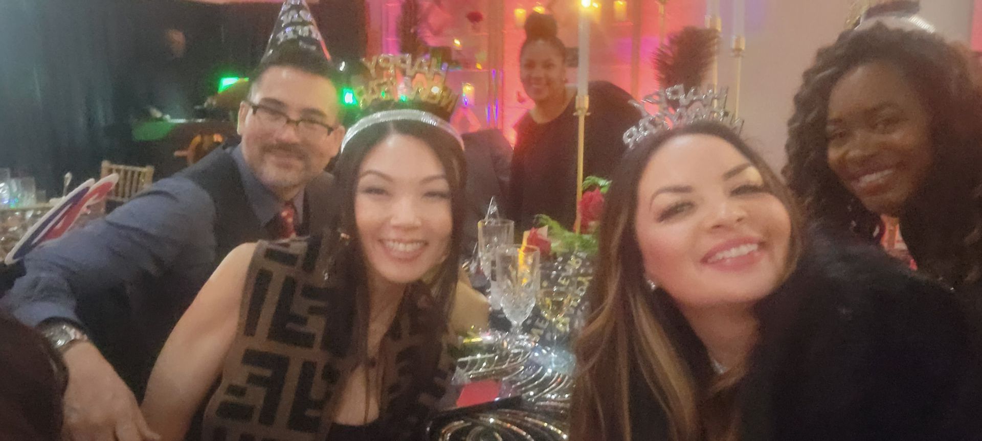 People smiling and posing for a photo at a party, likely New Year's Eve, with decorations and festive headwear.