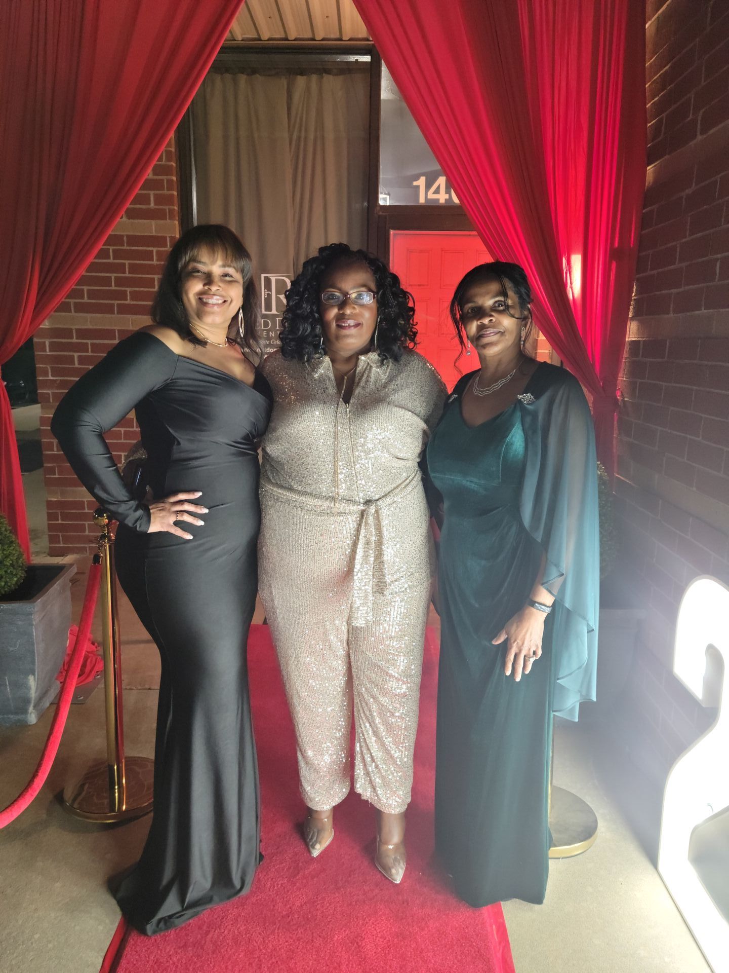 Three women in formal attire pose on a red carpet in front of a building entrance.