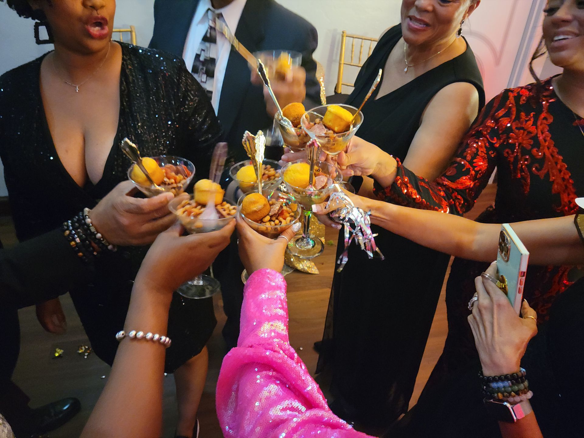 People clinking dessert-filled glasses at a party, cheerful expressions. Black dresses, pink accents, formal setting.