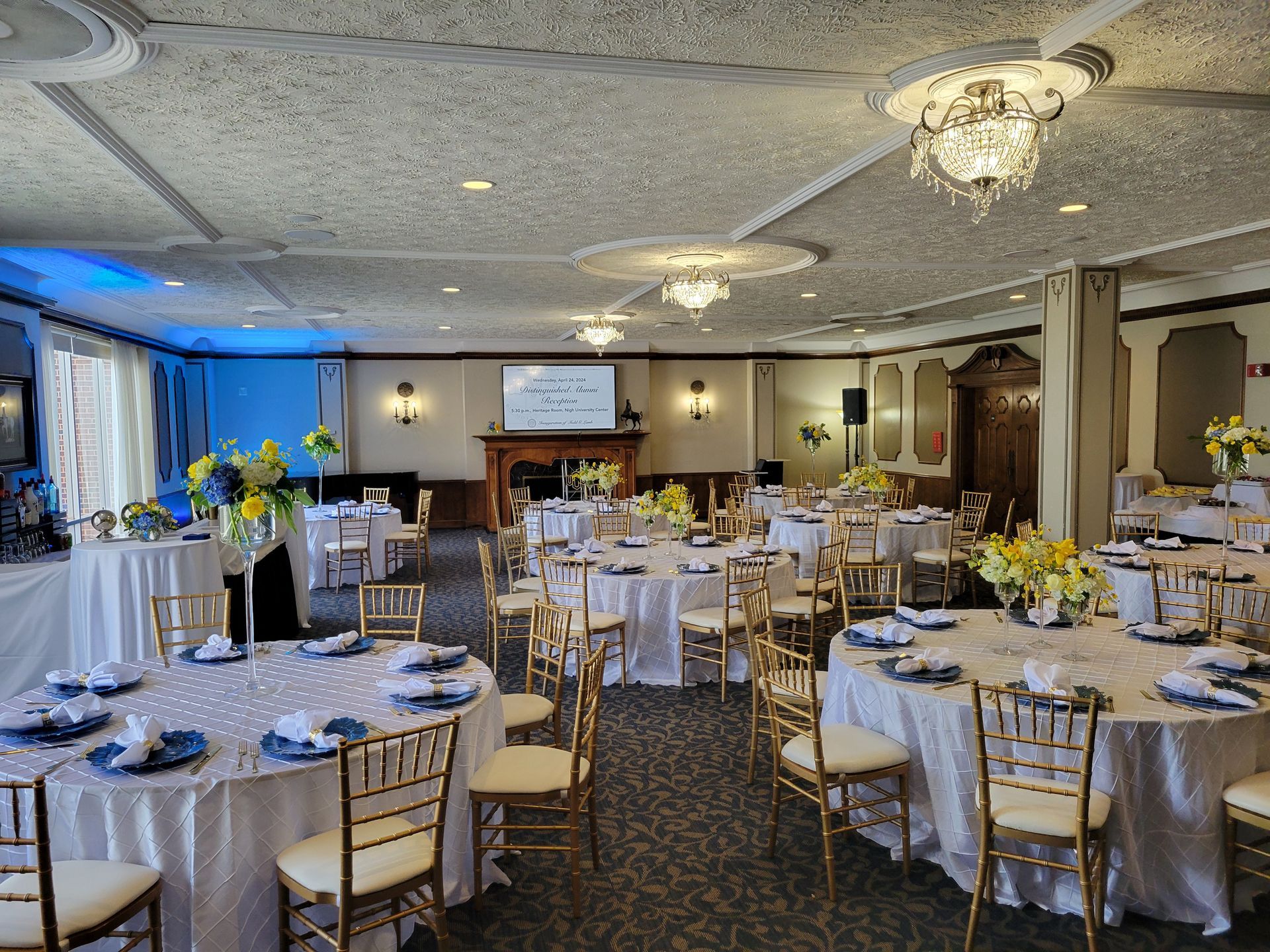 Formal banquet hall with round tables set for a celebration. Gold chairs, white tablecloths, and floral centerpieces.