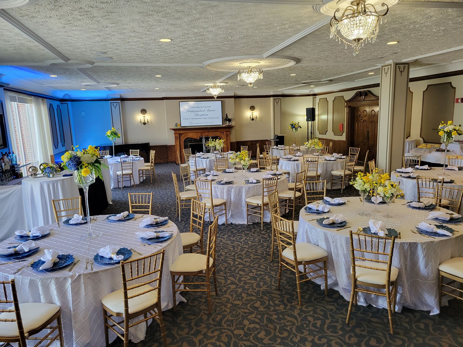 Reception hall with round tables set for a formal event. White linens, gold chairs, floral centerpieces.