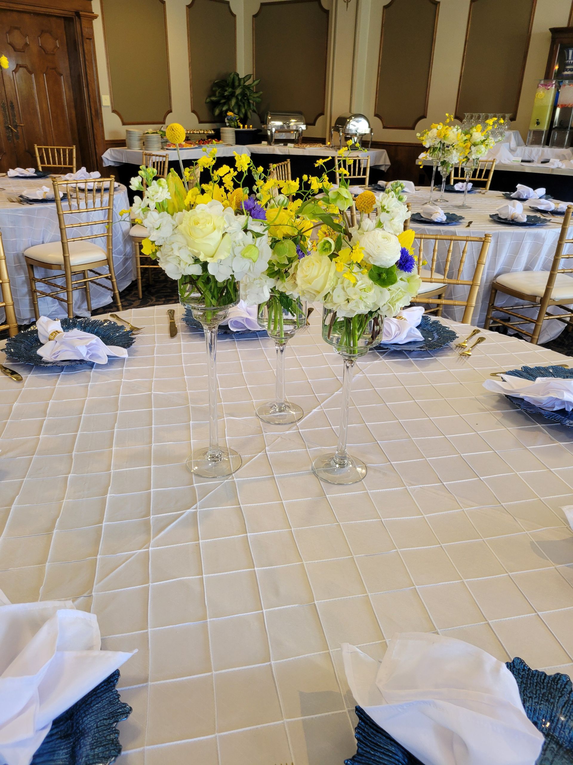 Elegant tables set for an event, featuring flower centerpieces in tall glasses. White tablecloths and gold chairs.