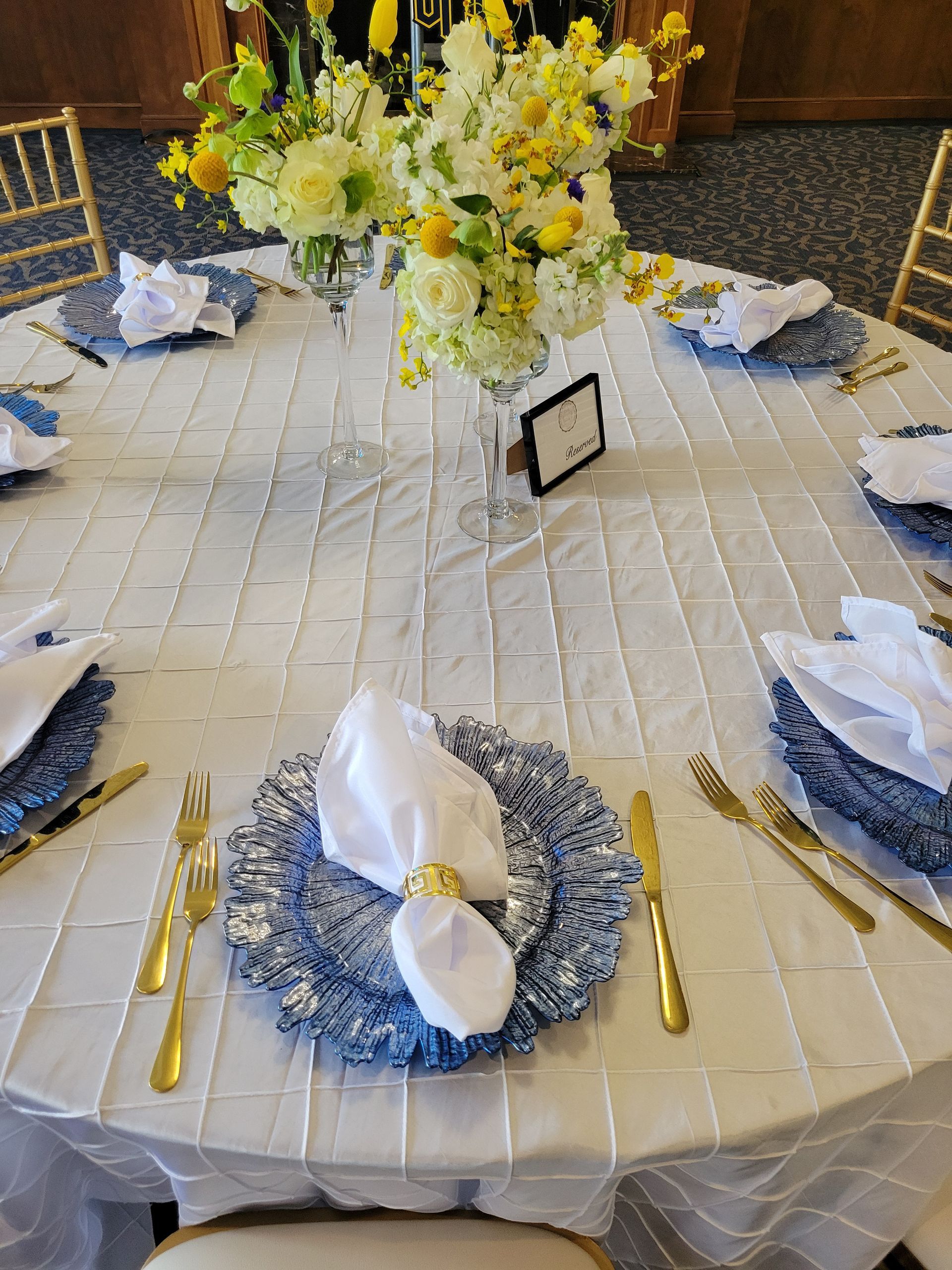 Round table set for a formal event with blue plates, gold utensils, white napkins, and a floral centerpiece.