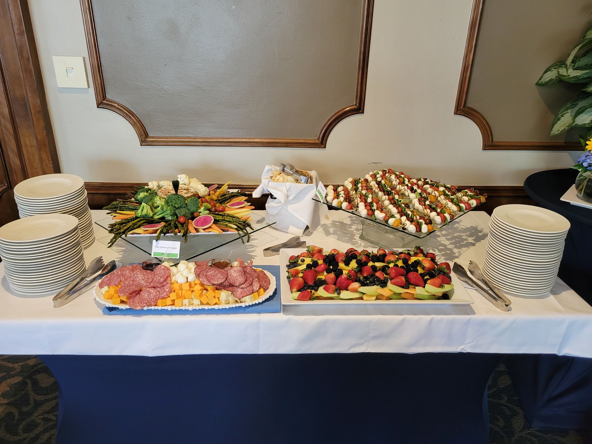 Buffet table with various foods: meats, veggies, fruit tarts, and egg bites. Includes plates, utensils, and serving tools.