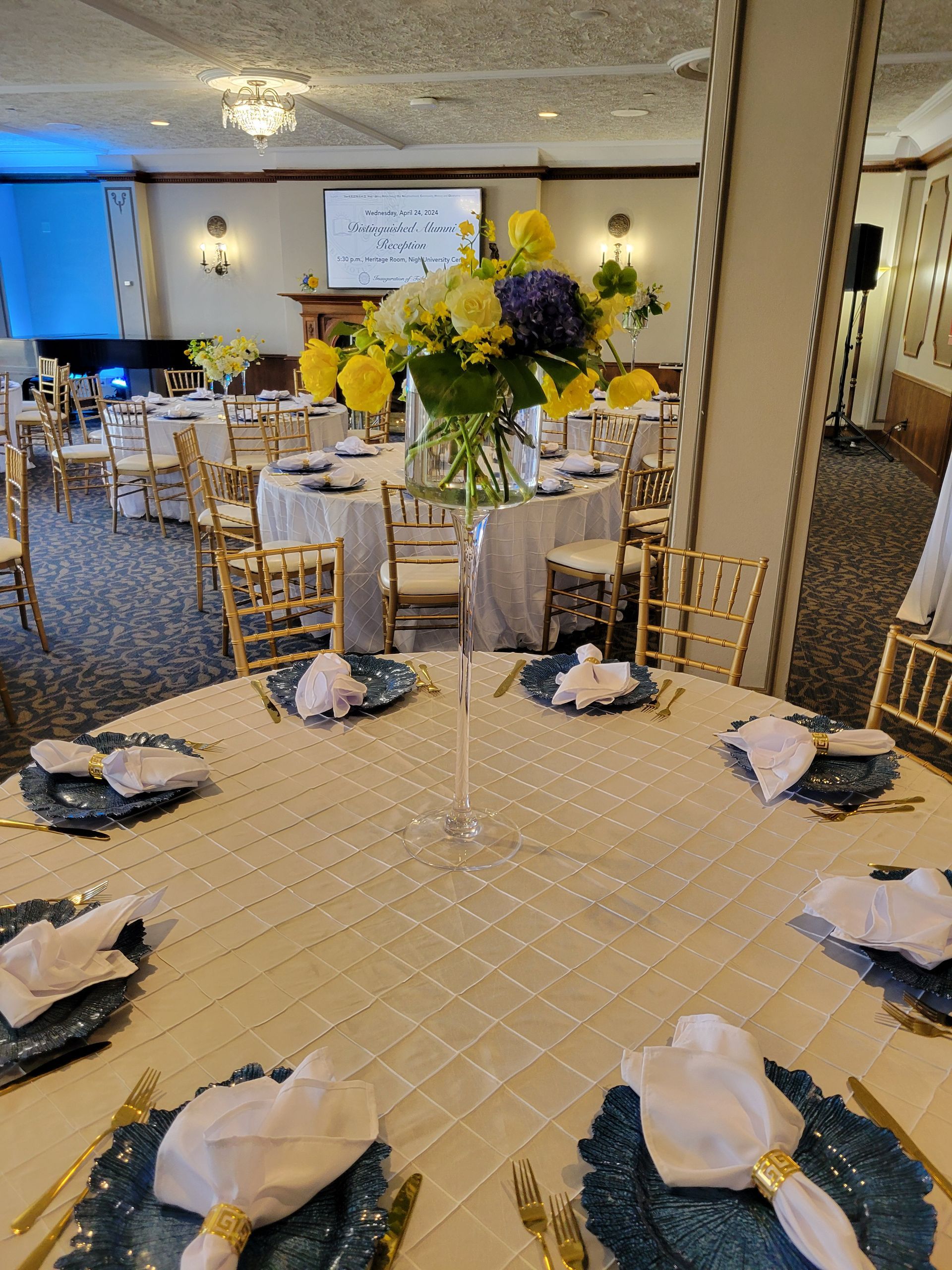 Round tables set for a celebration with floral centerpieces, blue chargers, and gold accents.