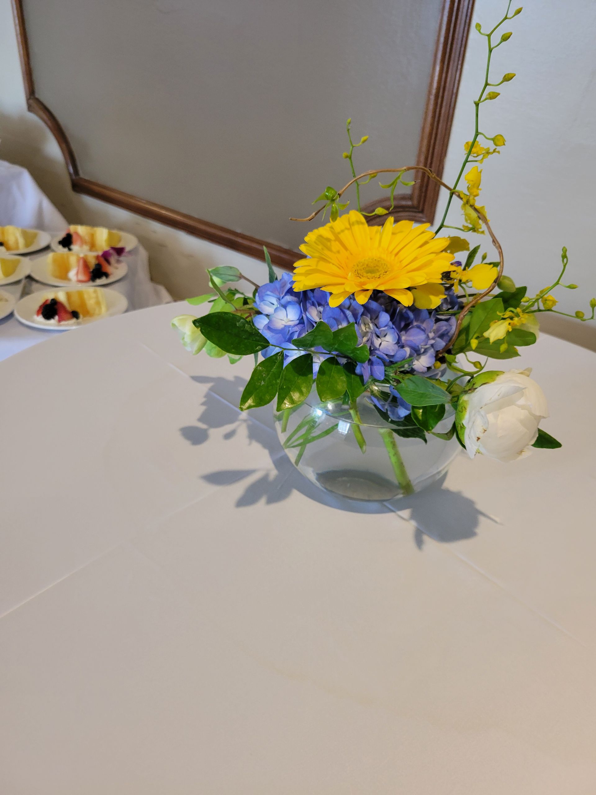 Floral arrangement with yellow gerbera daisy, blue hydrangeas, white rose, in a glass bowl on white tablecloth.