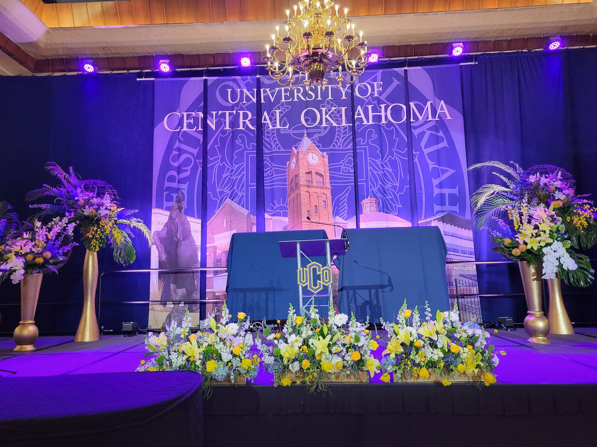 Stage setup at the University of Central Oklahoma with backdrop and floral arrangements.