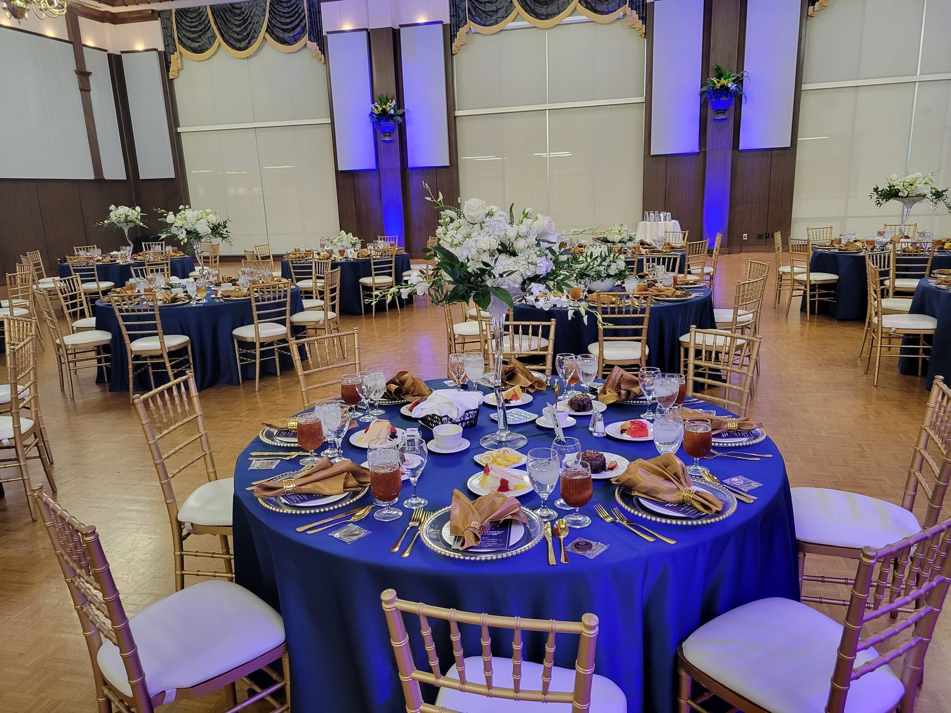 Round tables set for a formal event in a large room, with blue tablecloths, gold chairs, and floral centerpieces.
