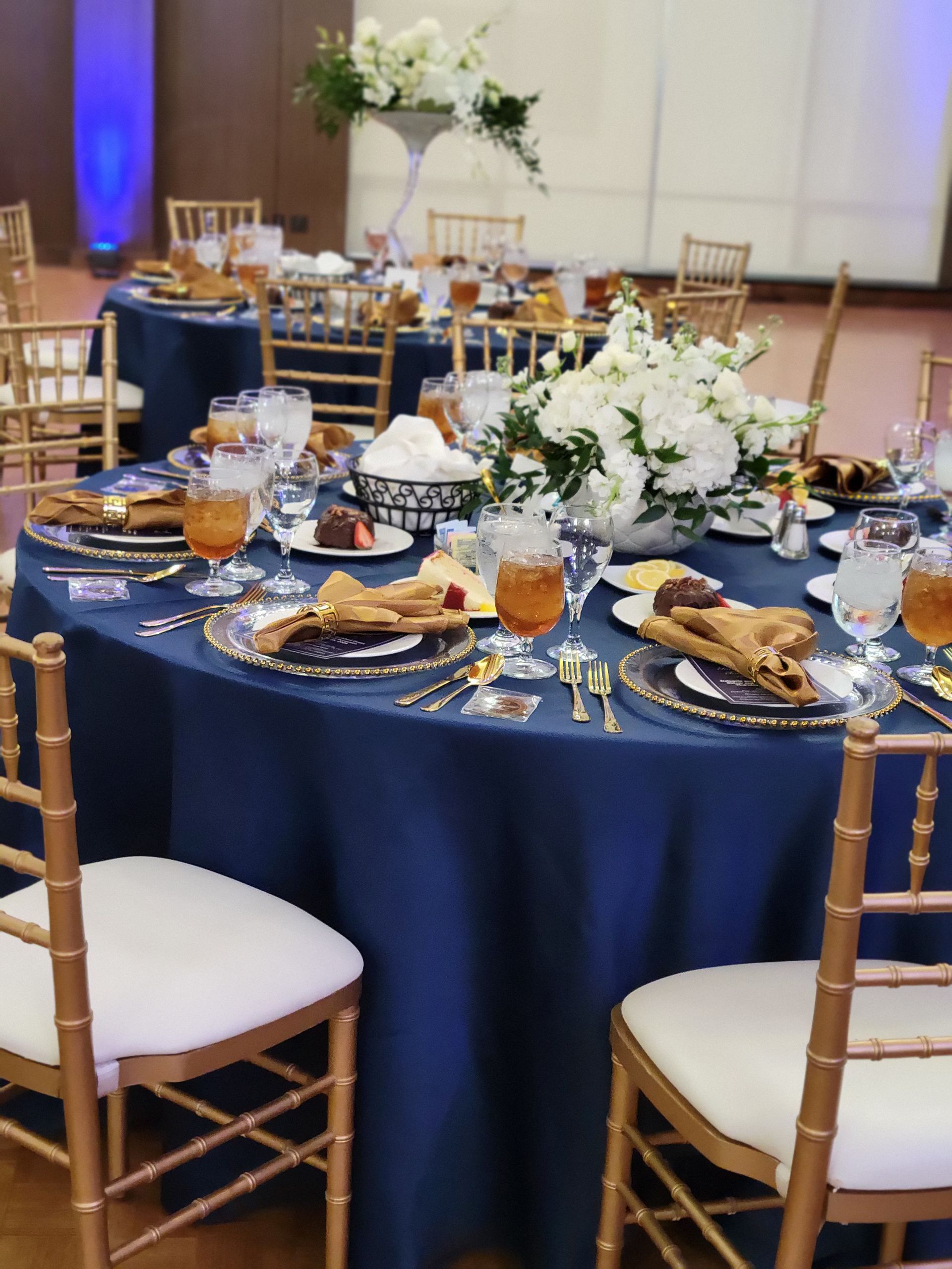 Round tables with blue tablecloths, gold chairs, and floral centerpieces, set for a formal event.