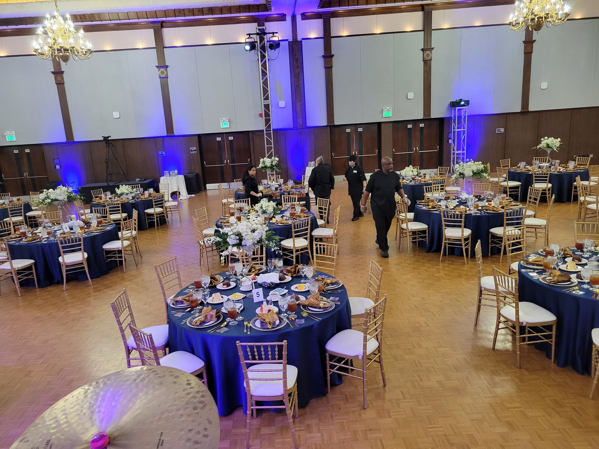 Large ballroom set for event; round tables with navy blue cloths, gold chairs, floral centerpieces.