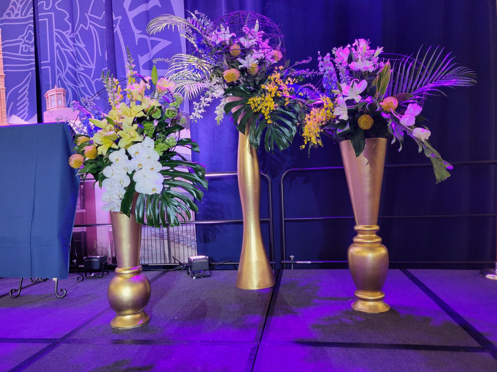 Three gold vases with colorful floral arrangements on a stage with a blue backdrop.